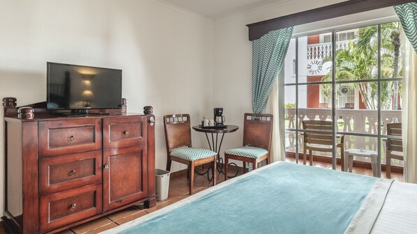 A hotel room with a TV on a wooden cabinet, two chairs with patterned cushions, a small table with a coffee maker, and a balcony with a view of palm trees. - Marien Puerto Plata - All Inclusive (Puerto Plata)