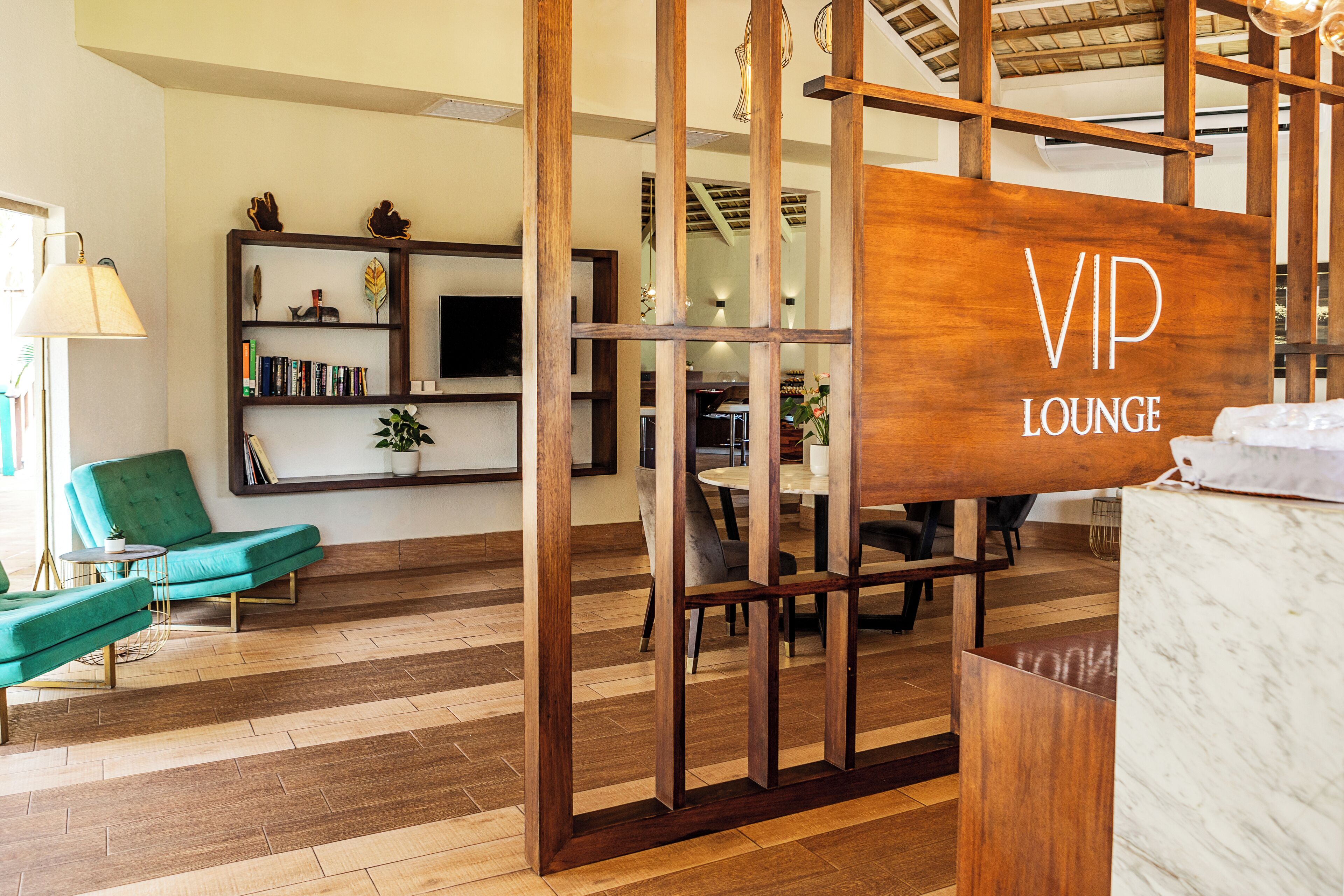 Een moderne lounge met een houten bord waarop 'VIP LOUNGE' staat, een groene fauteuil, een boekenkast en een eettafel met stoelen.