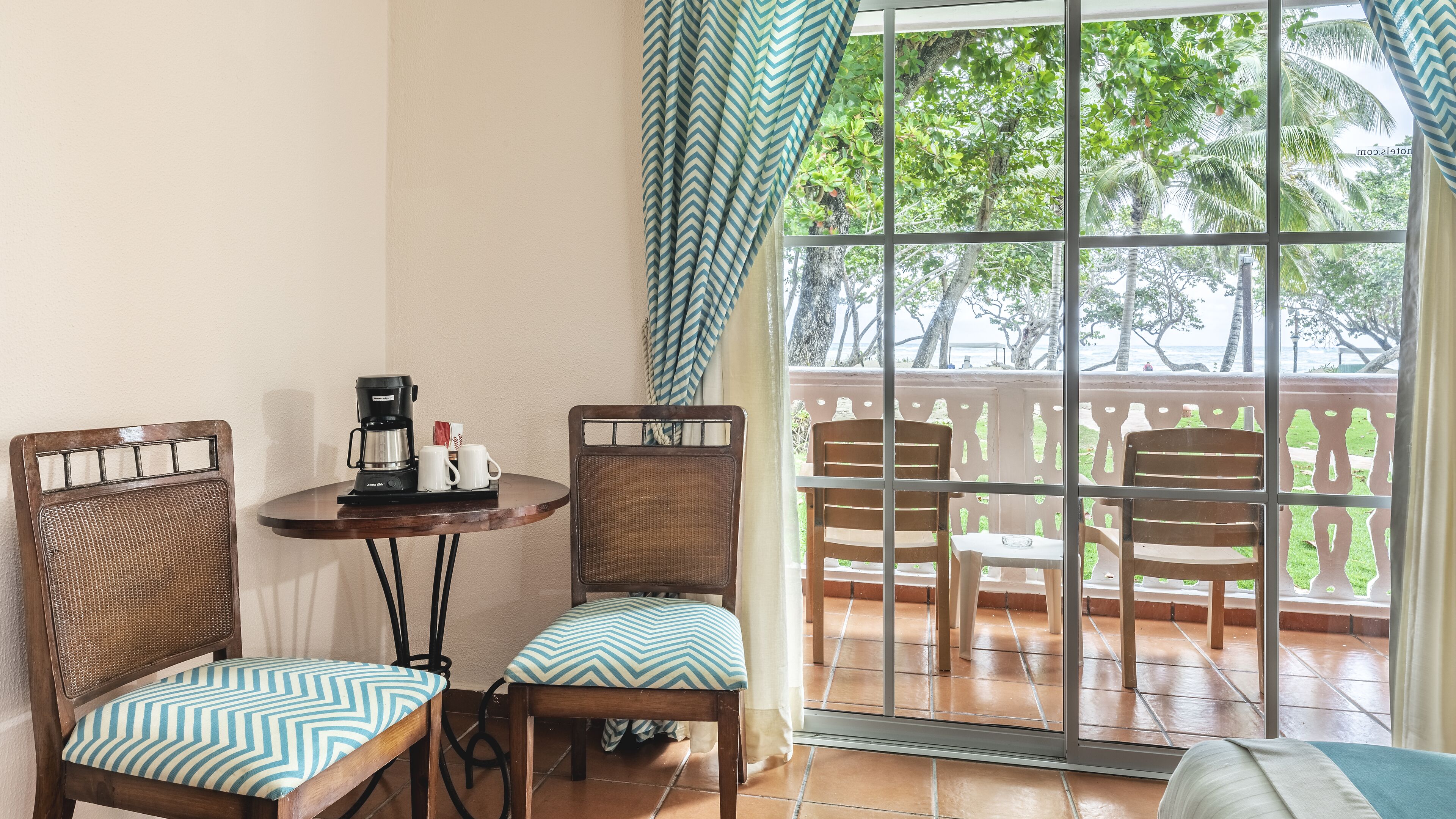 Ein kleines Zimmer mit Tisch, zwei Stühlen, einer Kaffeemaschine und Blick auf einen Balkon mit Sesseln und Geländer.