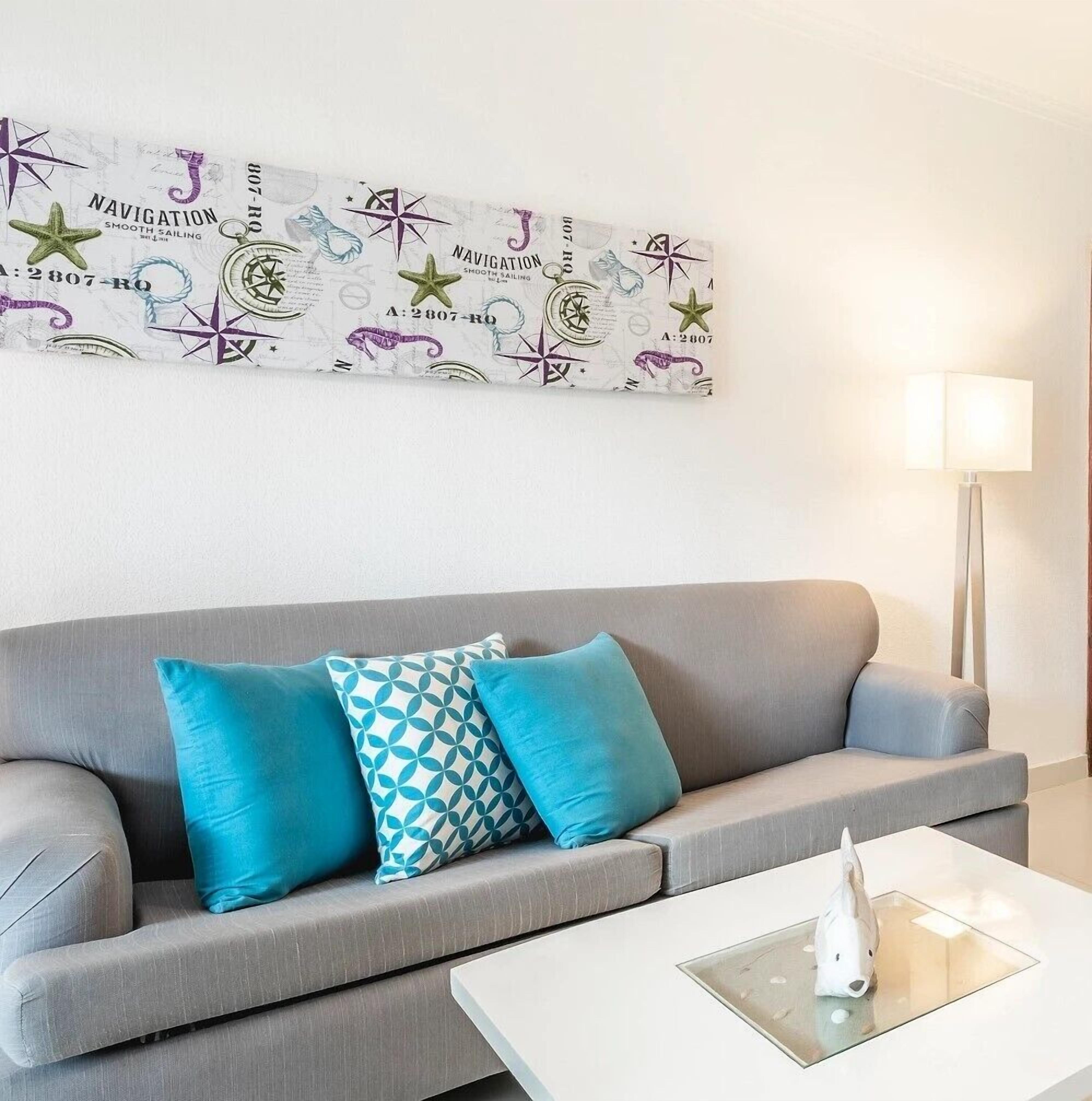 A modern living room with a grey sofa, two blue and one patterned teal pillow, a white coffee table with a white sculpture, and a wall-mounted lamp.