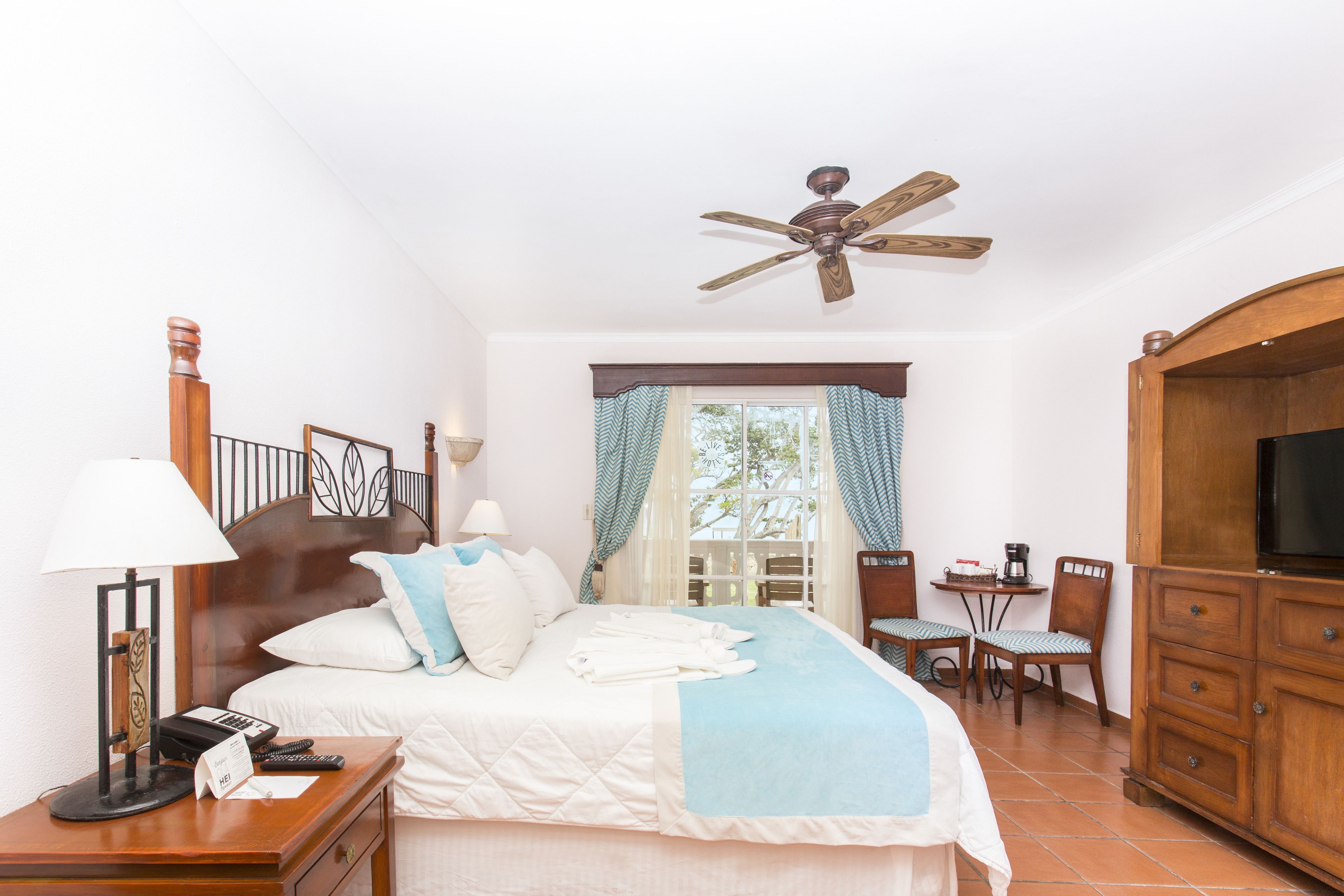 A hotel room with a bed, bedside table, lamp, ceiling fan, window with curtains, chairs, and a TV cabinet.