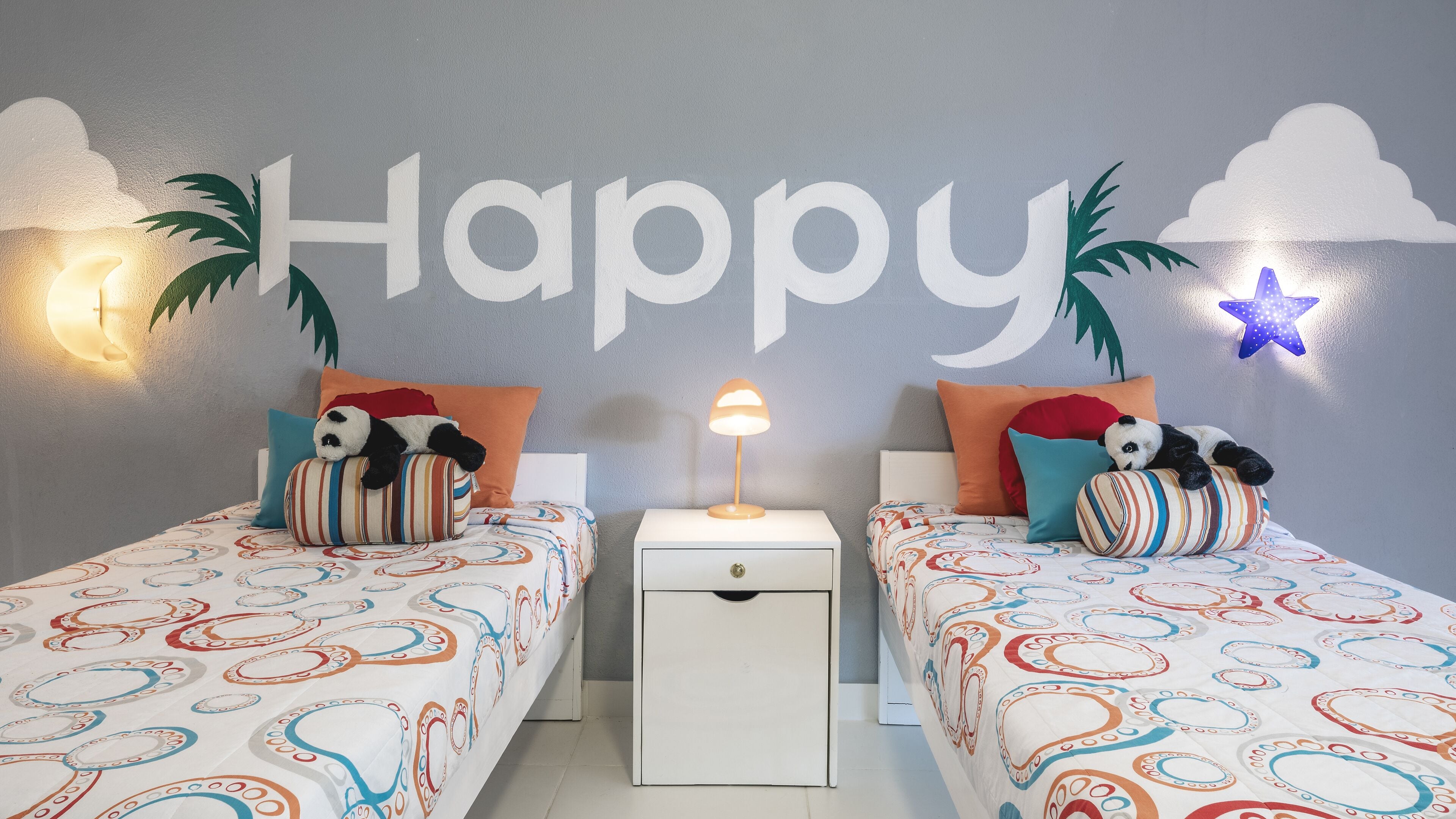 A child's bedroom with two beds, each with a panda plush toy, a nightstand with a lamp, and a wall with 'Happy' and cloud decorations.