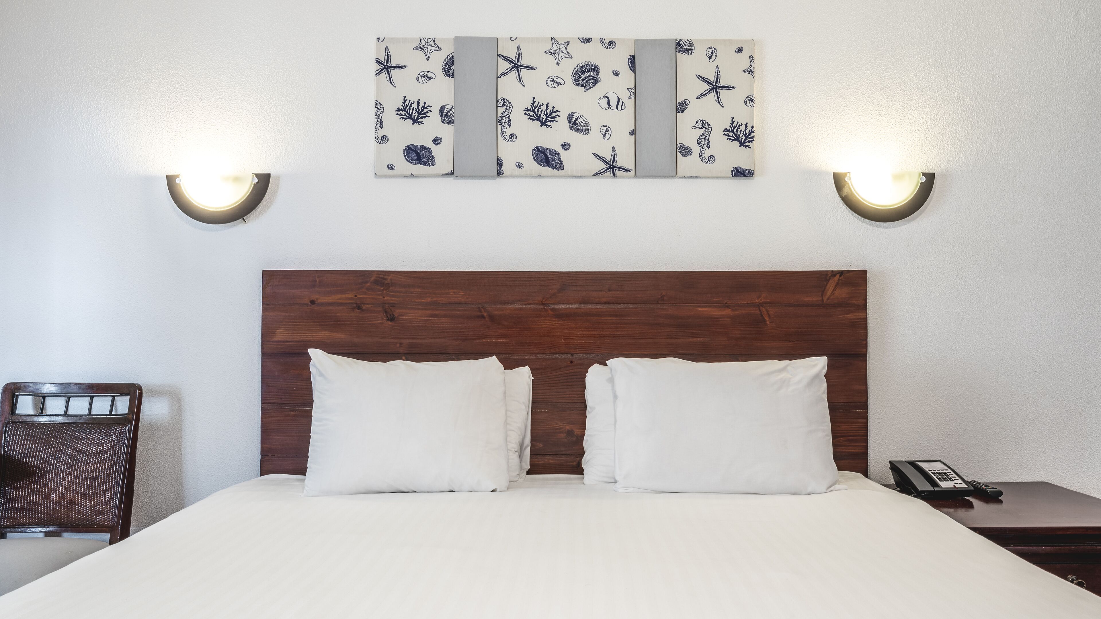 Habitación de hotel con cabecera de madera, dos almohadas blancas, cama, mesita de noche con teléfono e iluminación en la pared.