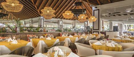 Un restaurant doté de nombreuses tables, aménagé pour accueillir les clients, avec un décor tropical caractérisé par des plafonds en chaume et des meubles en osier.