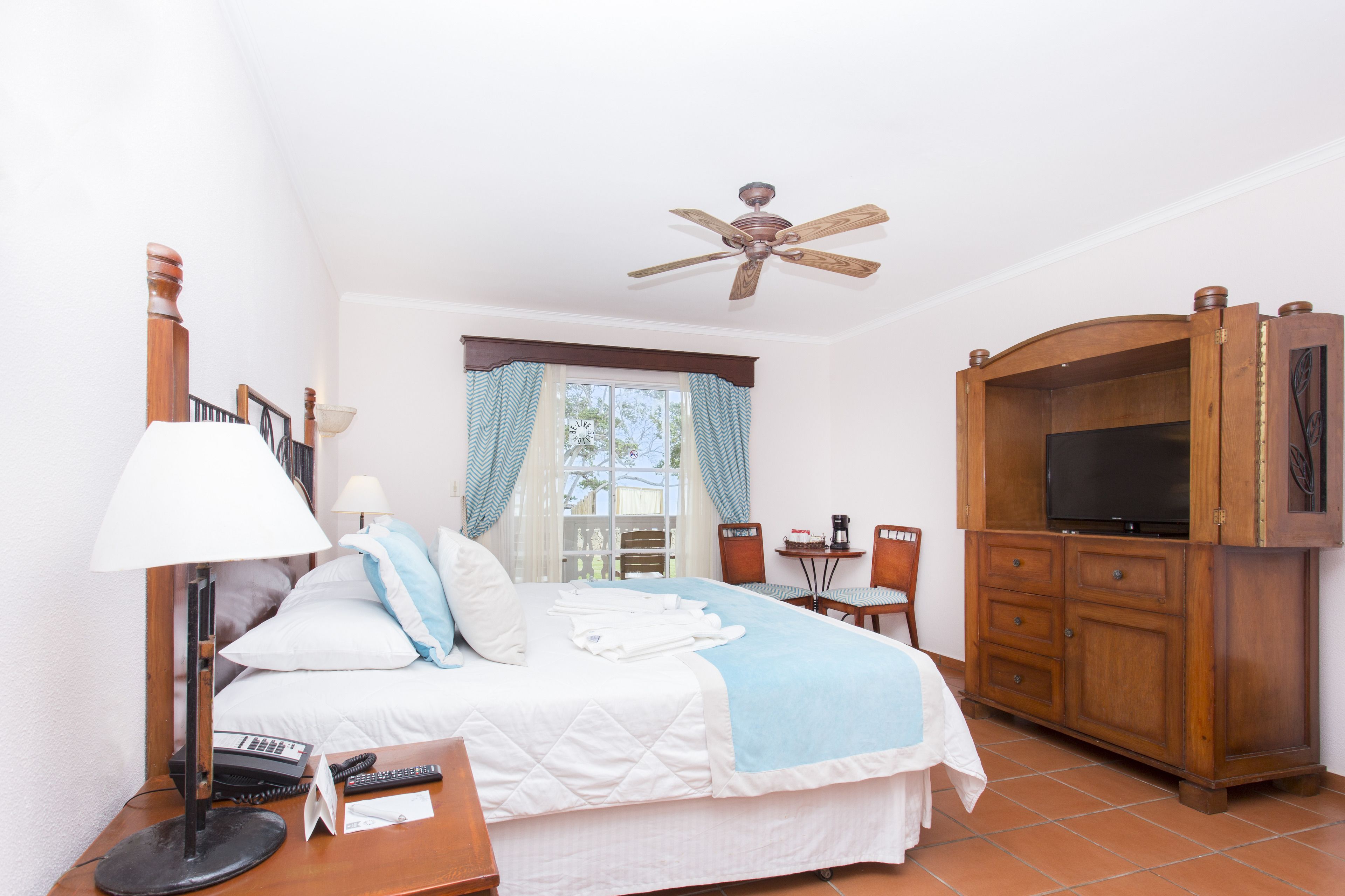 A hotel room with a bed, a desk with a lamp, a TV cabinet, and a ceiling fan.