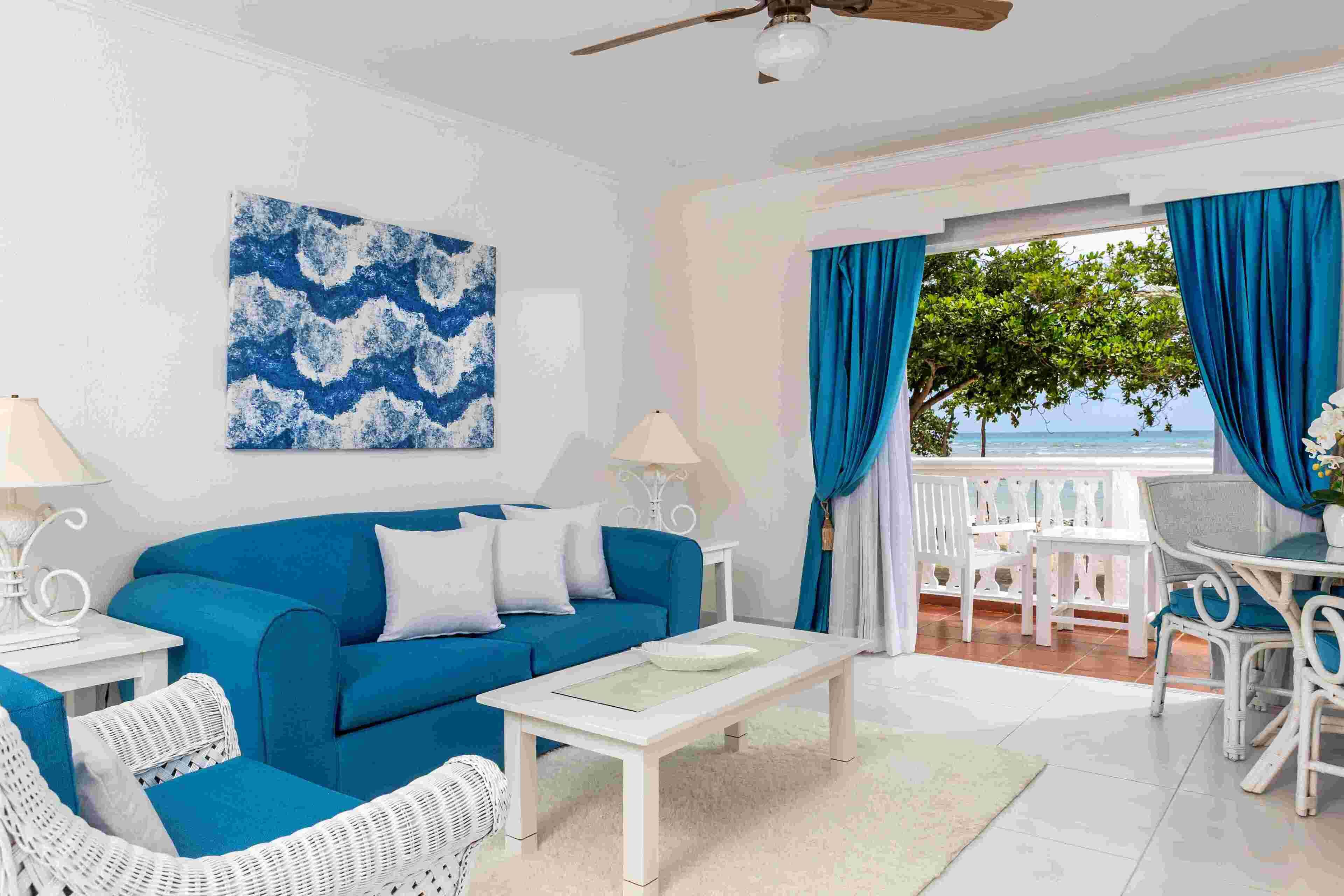 Ein Wohnzimmer mit einem blauen Sofa, weißen Sessel und einem weißen Couchtisch, mit Blick auf das Meer vom Balkon aus.