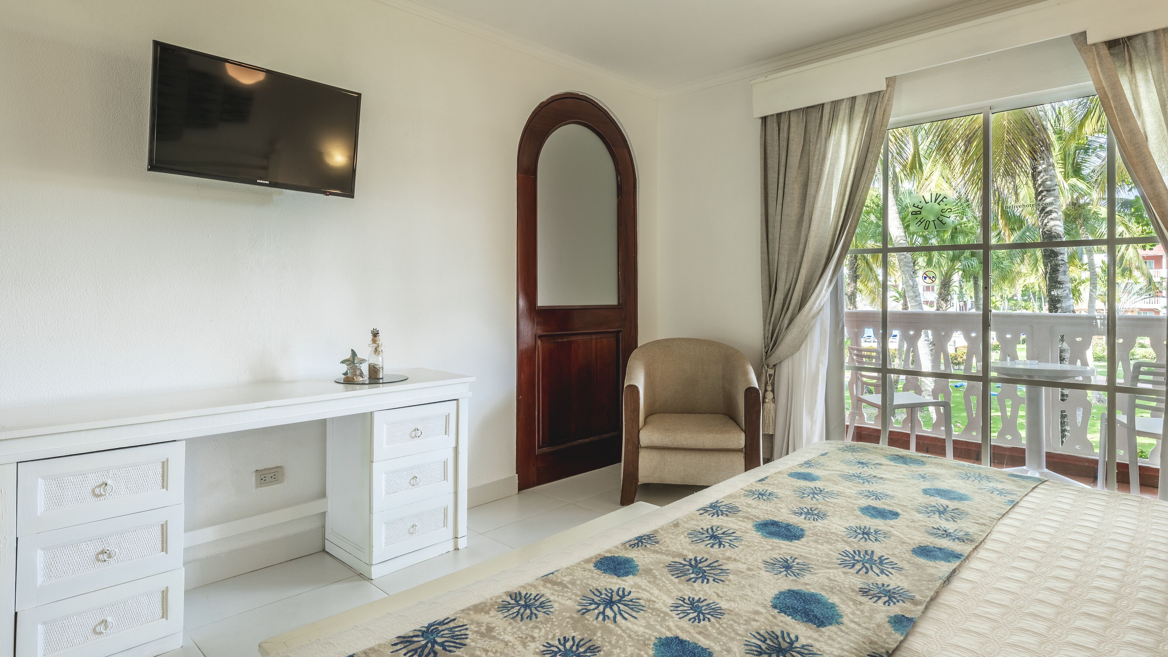 Un dormitorio con cama, escritorio con cajones, silla, televisor montado en la pared y ventana con cortinas.