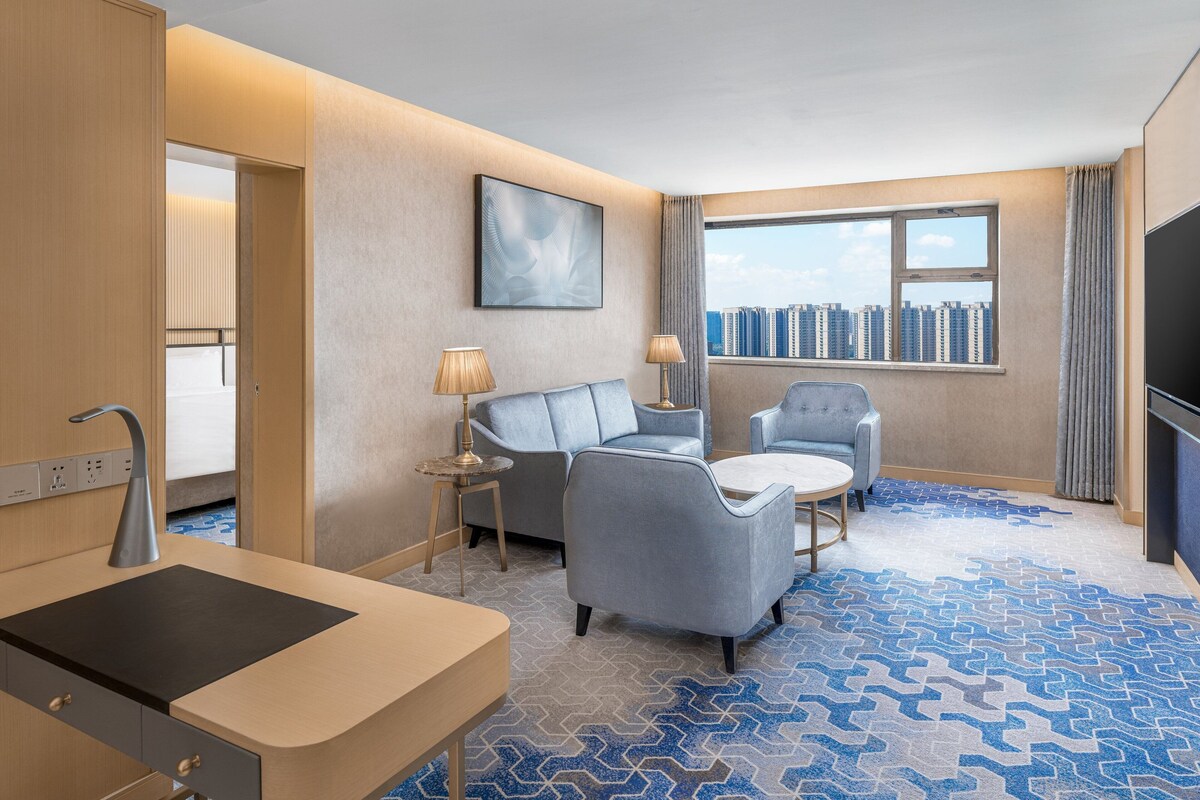 Ein Hotelzimmer im modernen Stil, mit einem großen Bett, einem Schreibtisch mit Spüle, zwei Sesseln, einem kleinen runden Tisch und einem großen Fenster mit Blick auf die Stadt.
