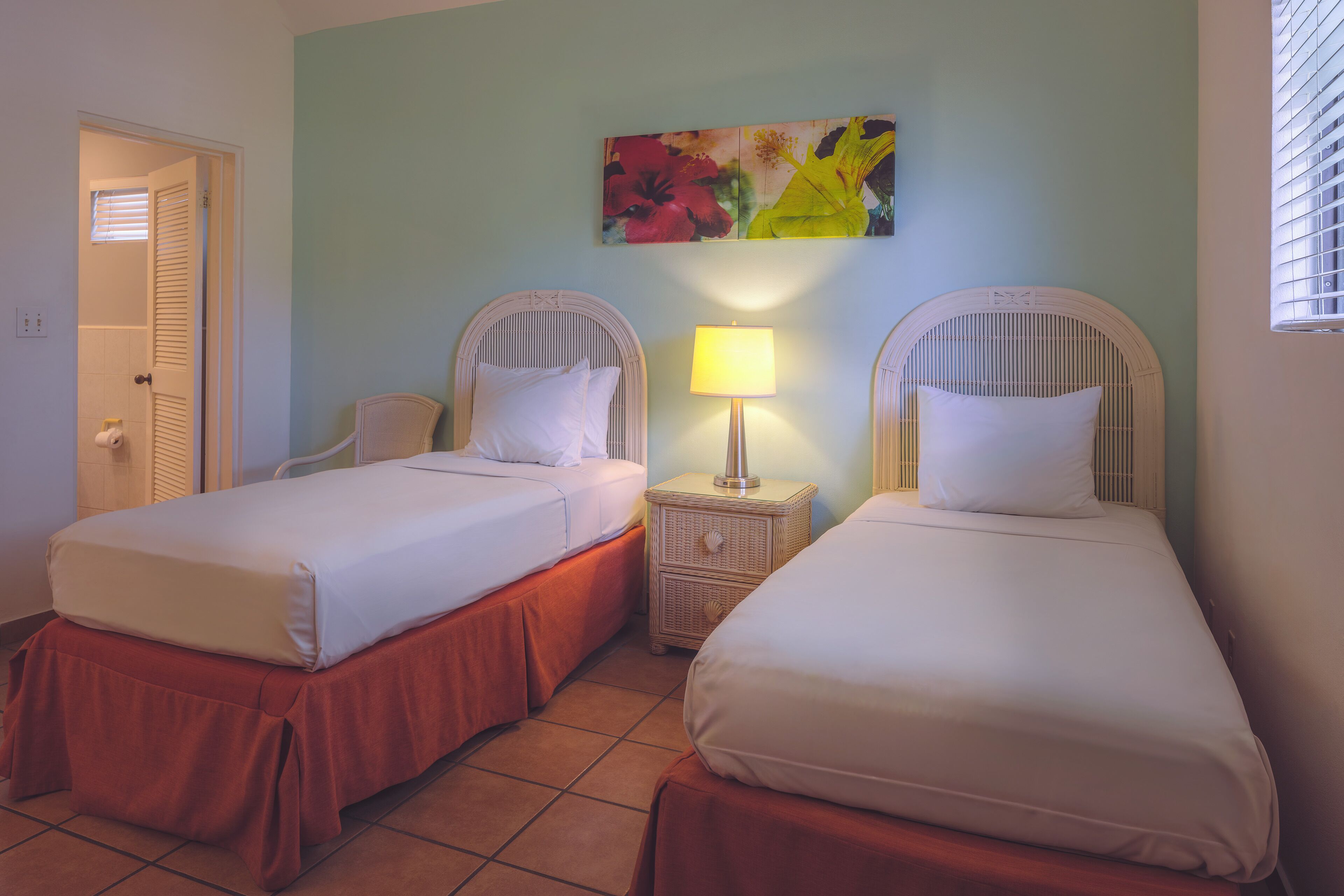 A hotel room with two single beds, a small table with a lamp, a chair, and a painting on the wall.