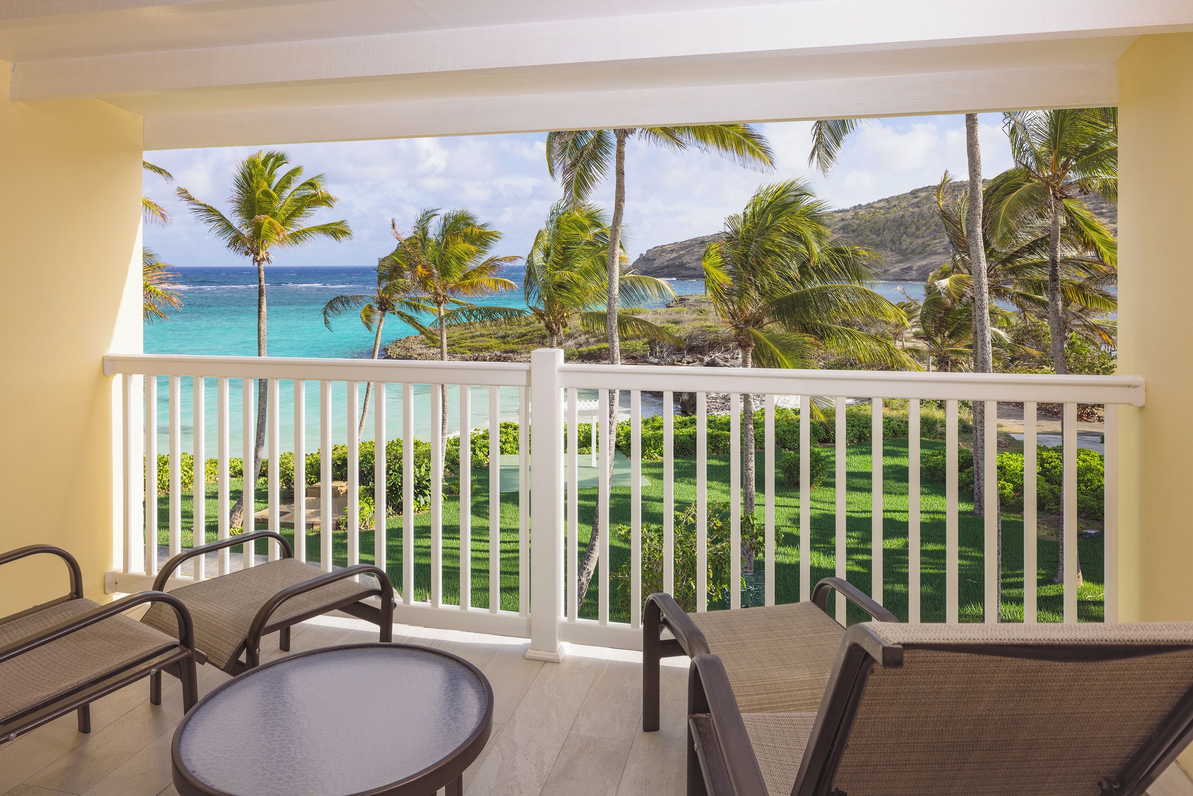A balcony with a view of the ocean and palm trees, furnished with chairs and a table.