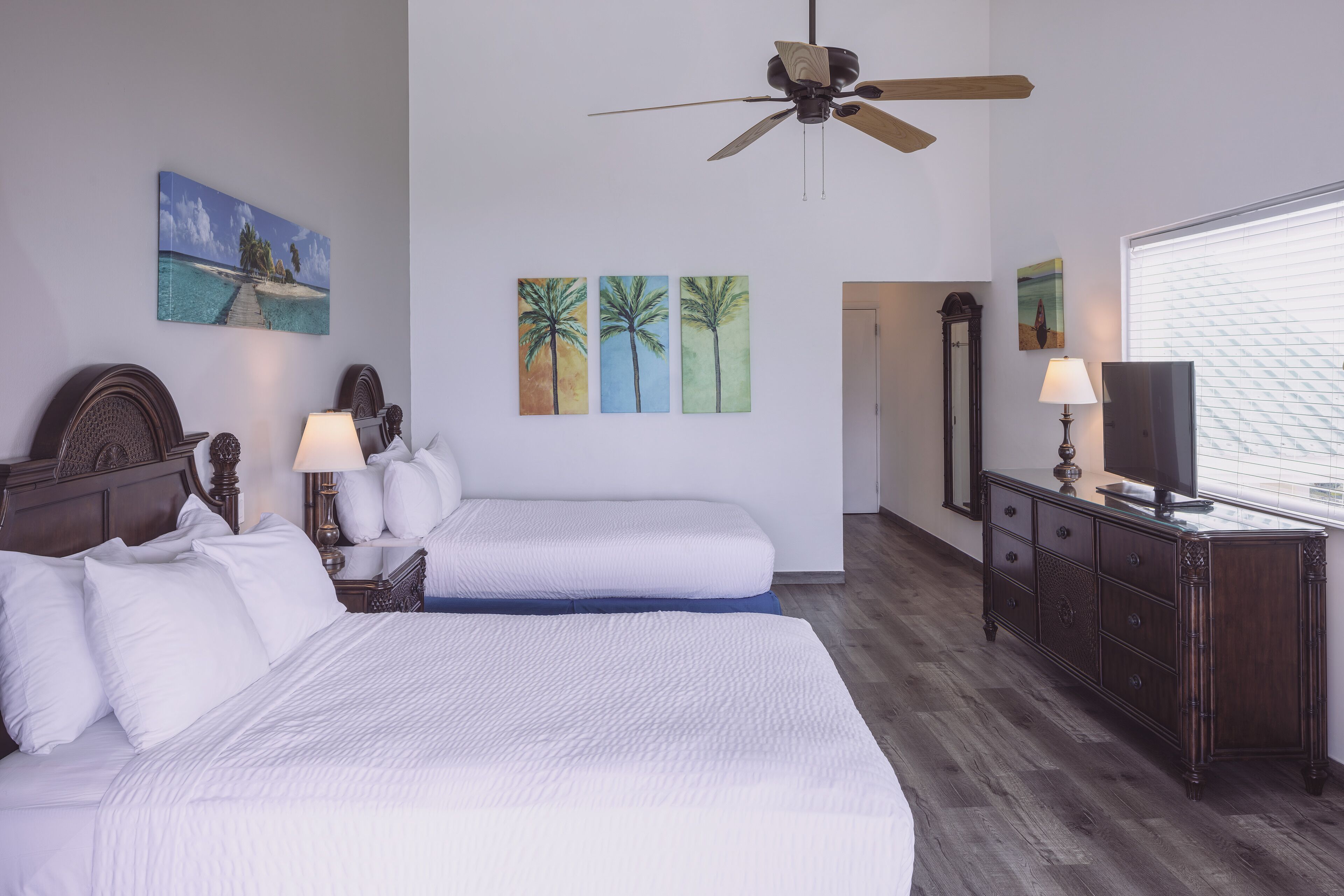 A hotel room with two beds, a ceiling fan, artwork on the walls, a dresser with a TV, and a window with blinds.