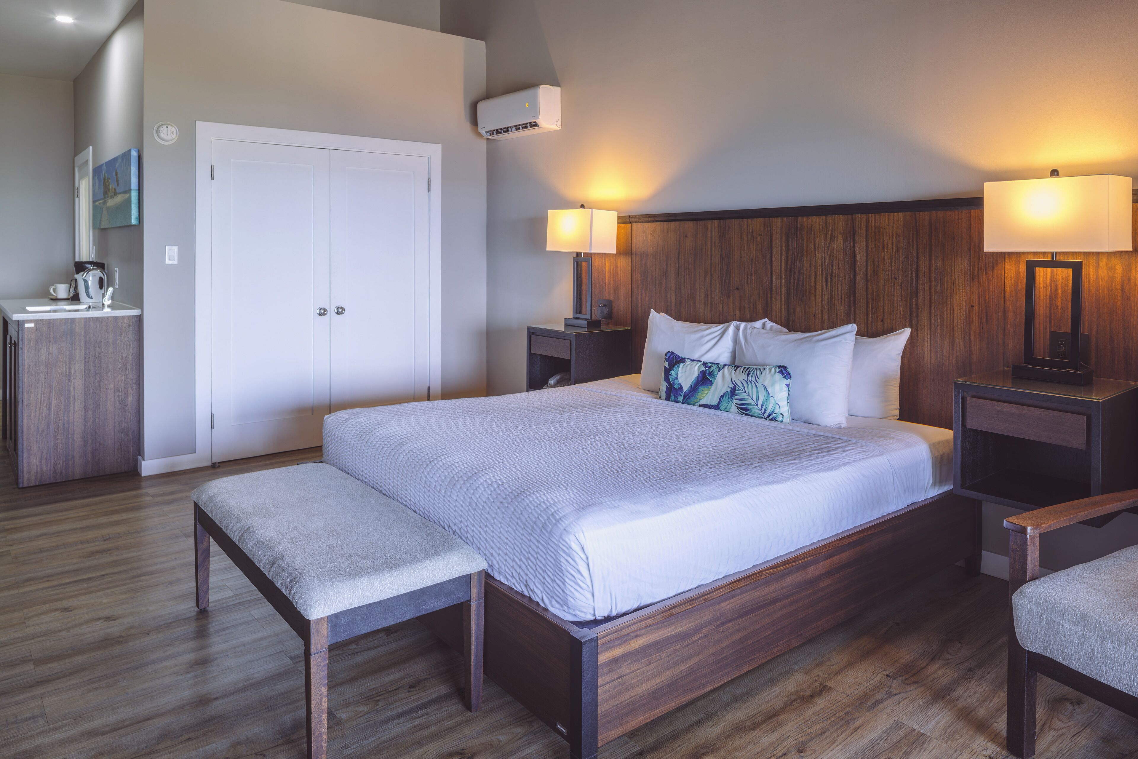 A hotel room with a large bed, bedside lamps, a wooden headboard, a bench, a wardrobe, and an air conditioning unit.