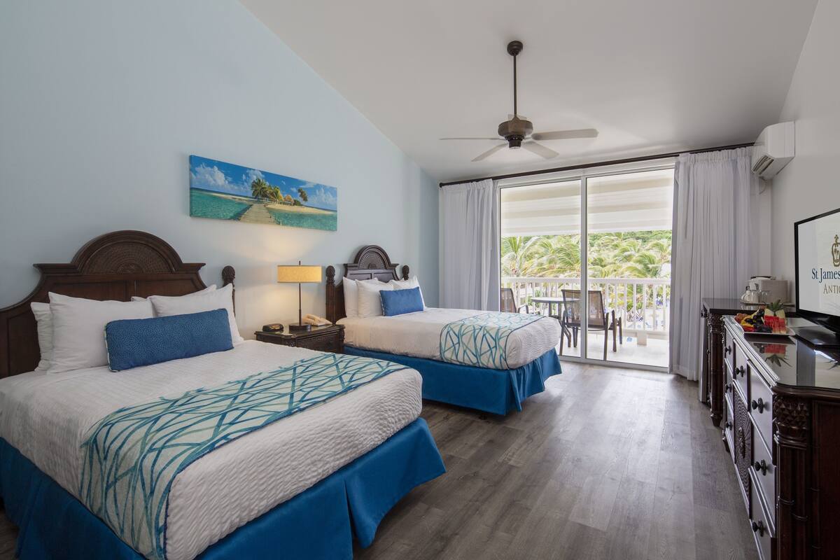Ein Hotelzimmer mit zwei Betten, einem Deckenventilator, einem Strandbild, einem Balkon mit Blick auf Palmen und einer Minibar.