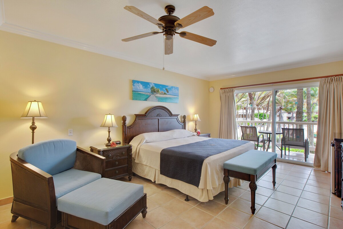 A bedroom with a bed, a chair, a table, a ceiling fan, and a window with curtains.