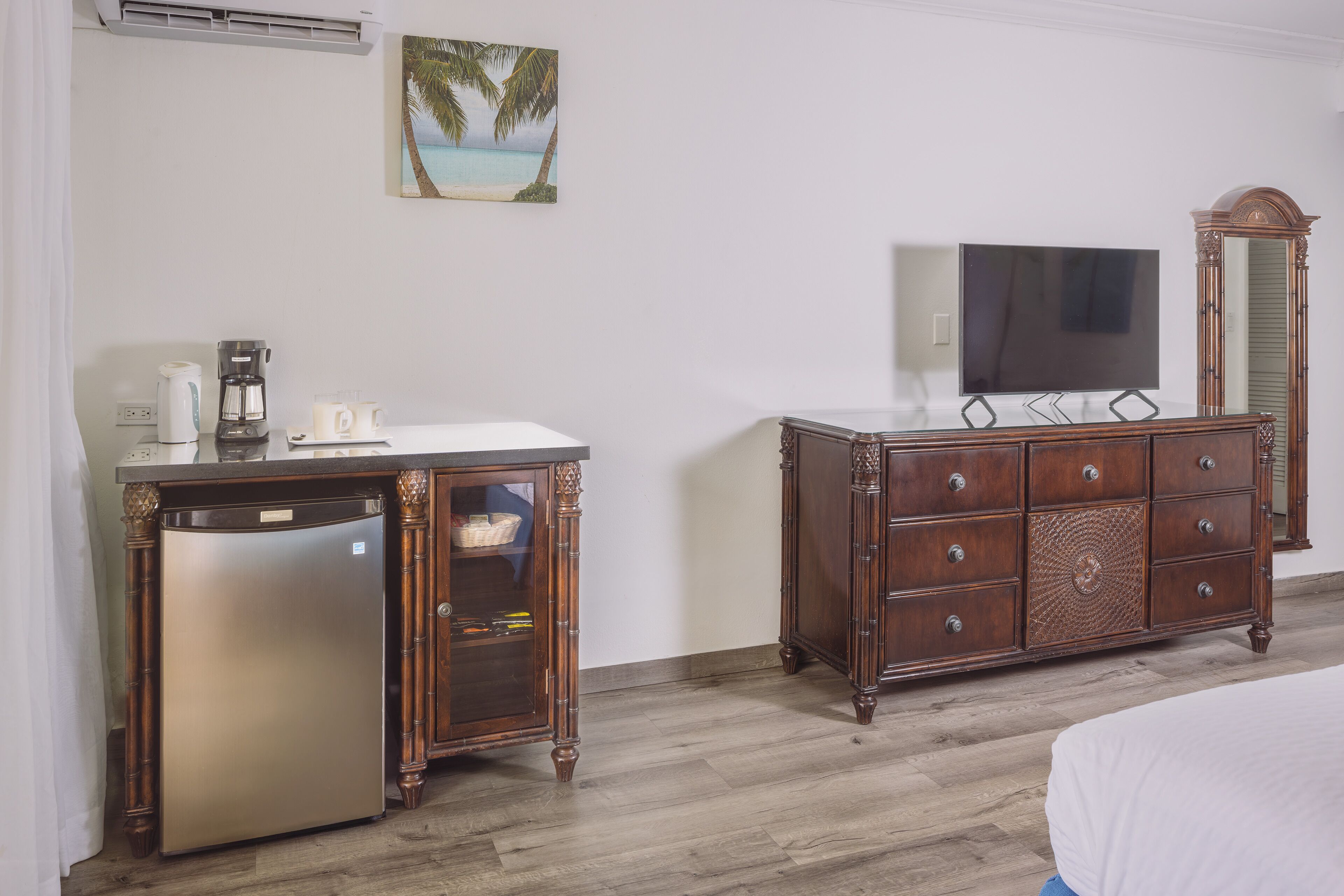 A hotel room with a bed, a wooden dresser, a refrigerator, a coffee maker, a television, and a wall-mounted air conditioner.