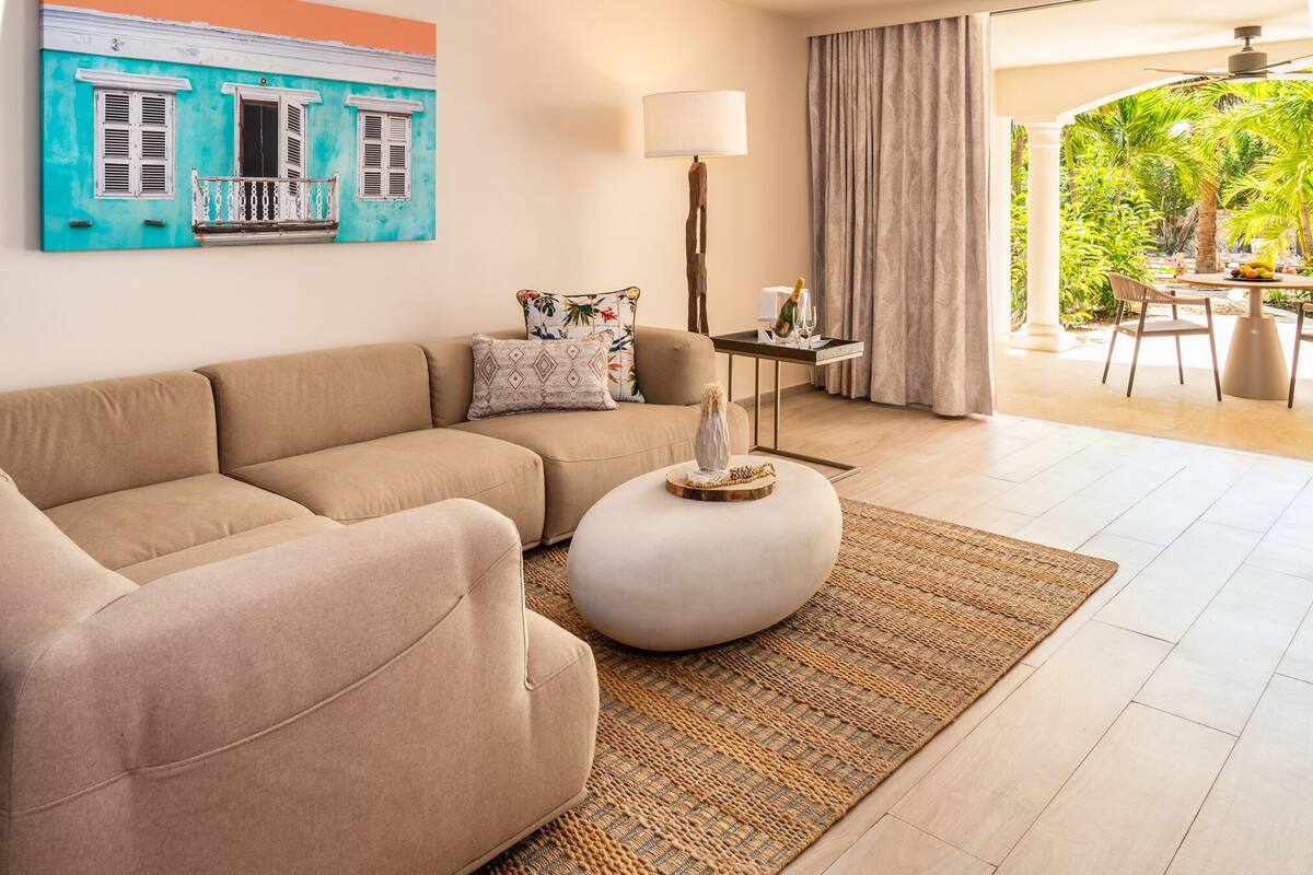 A living room with a beige sofa, a round coffee table, and a painting of a blue building. There is a lamp, a small table with a bottle, and a view of a garden through a window.