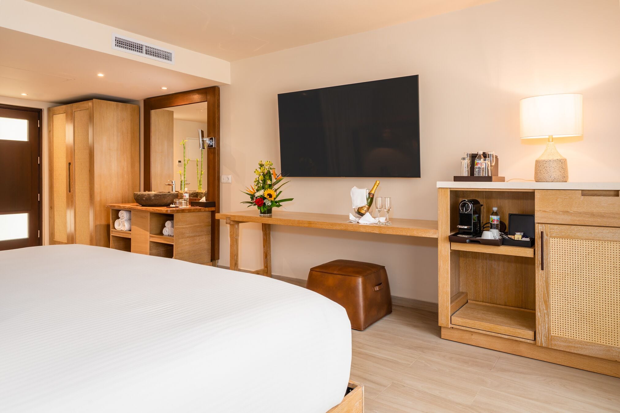 Quarto de hotel com uma cama grande, guarda-roupa de madeira, uma mesa com luminária, televisão de tela plana e um banquinho pequeno.