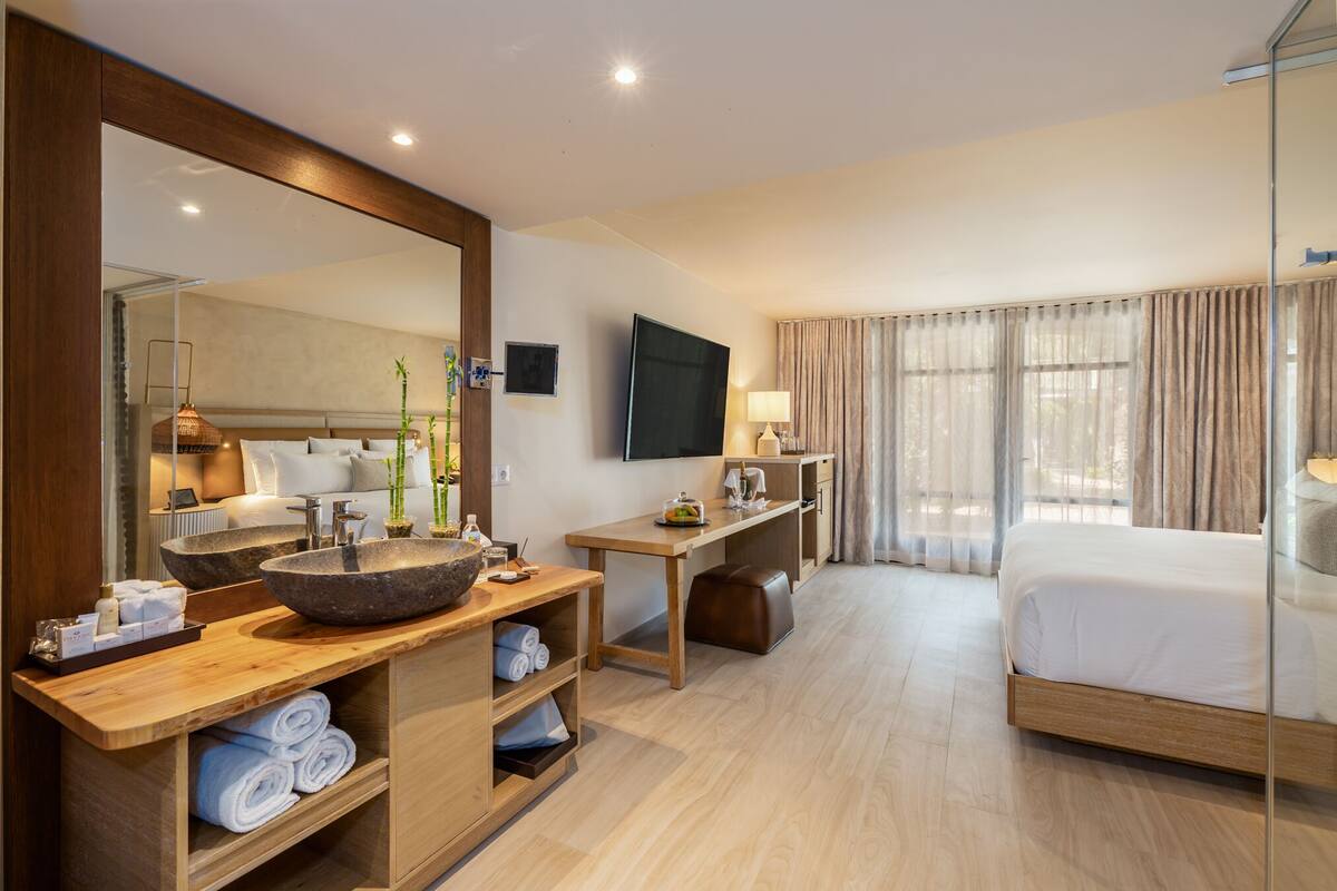 A modern hotel room with a large bed, a wooden vanity with a large sink, a flat-screen TV, and a large mirror.