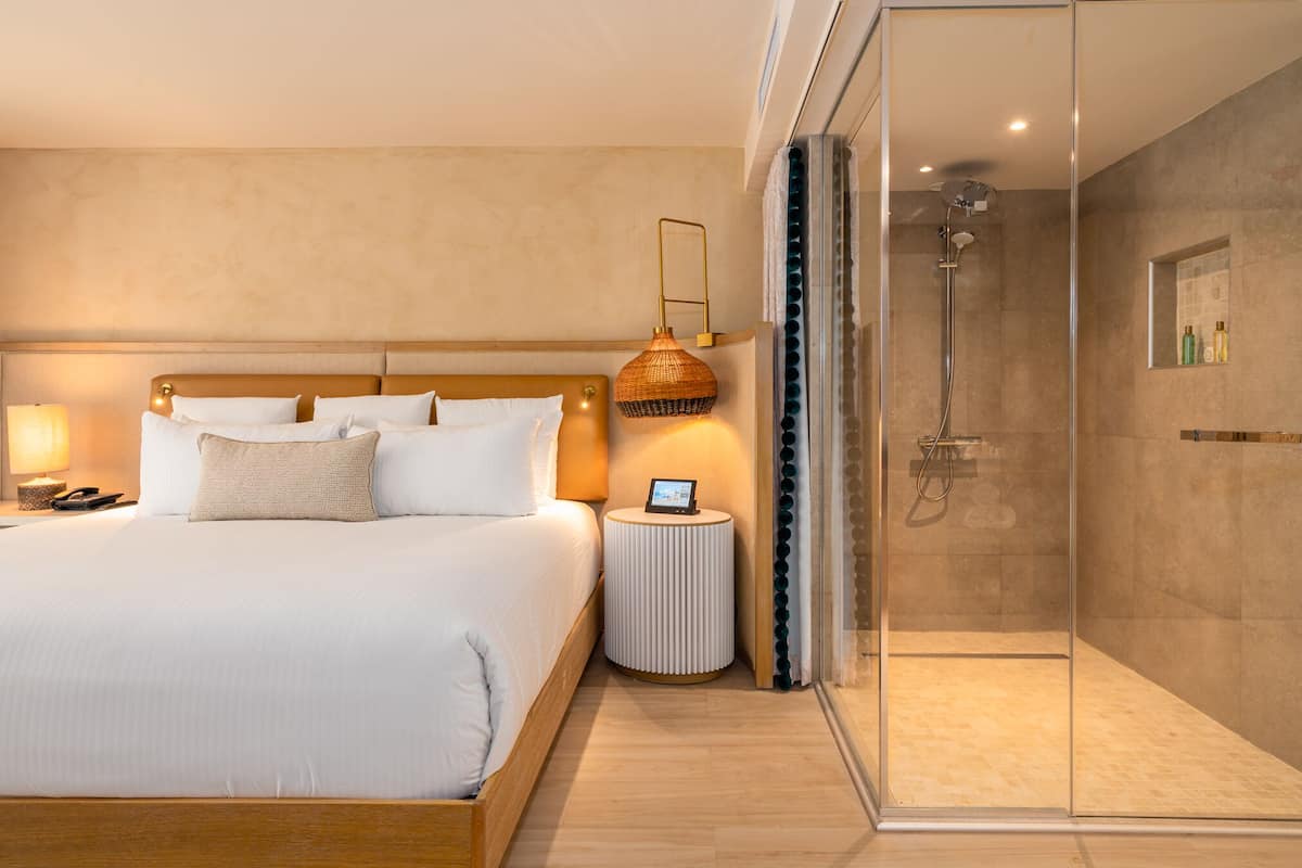 A modern hotel room with a large bed, bedside table, lamp, and a glass-enclosed shower area.