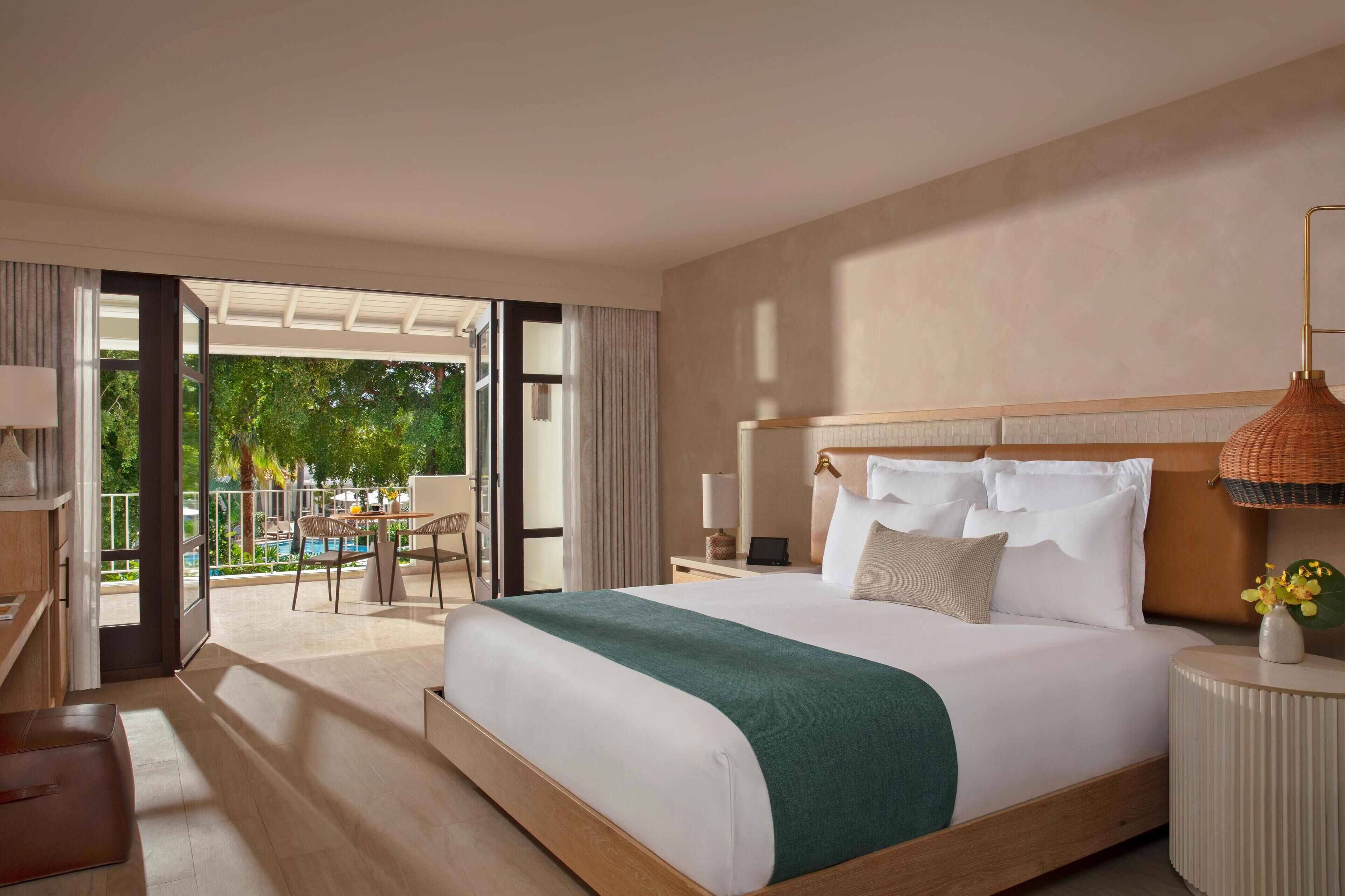 Een hotelkamer met een groot bed, een nachtkastje, een lamp en uitzicht op een buitenzwembad.