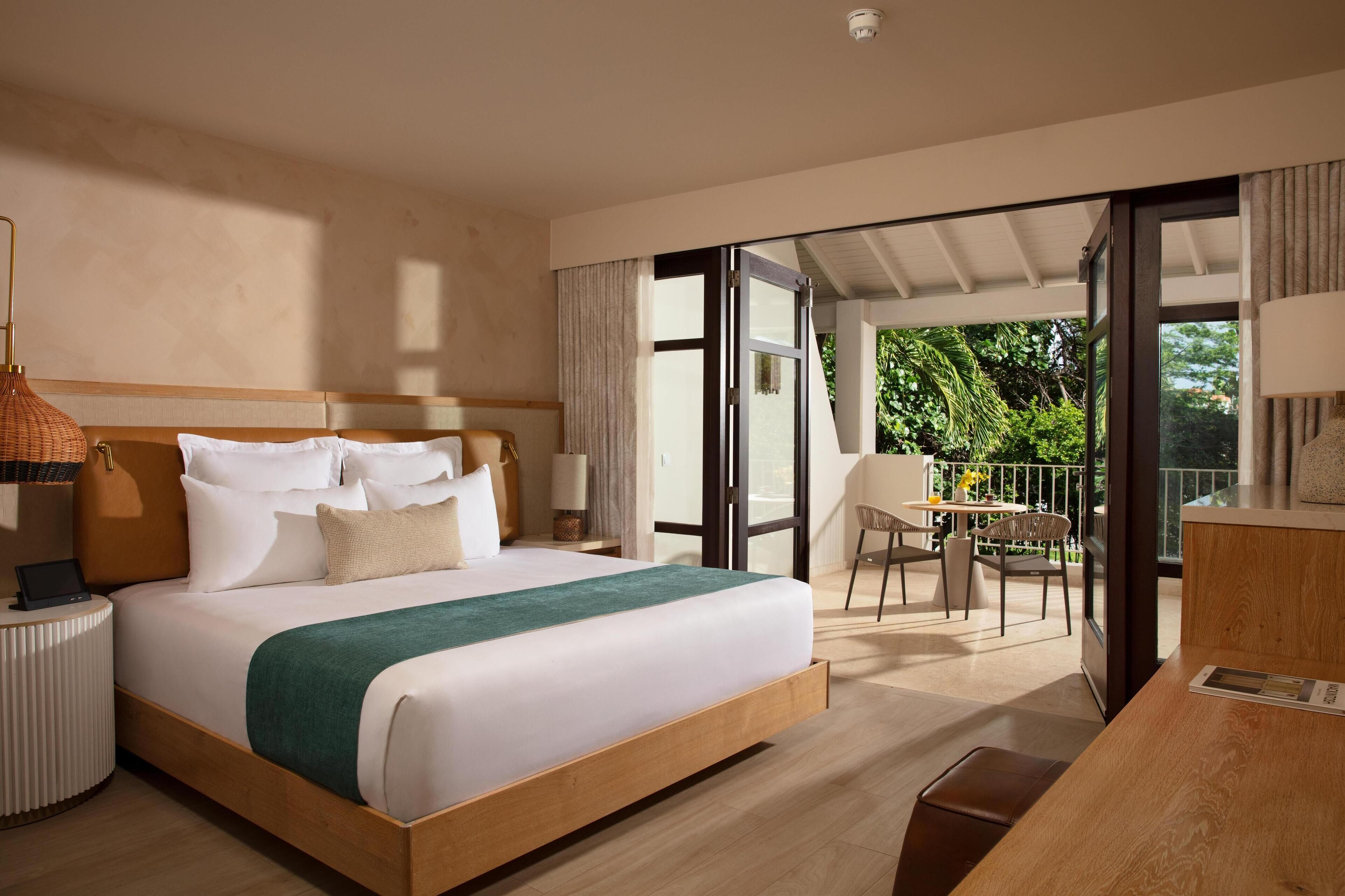 A modern hotel room with a large bed, a desk, and a balcony with a view of greenery.