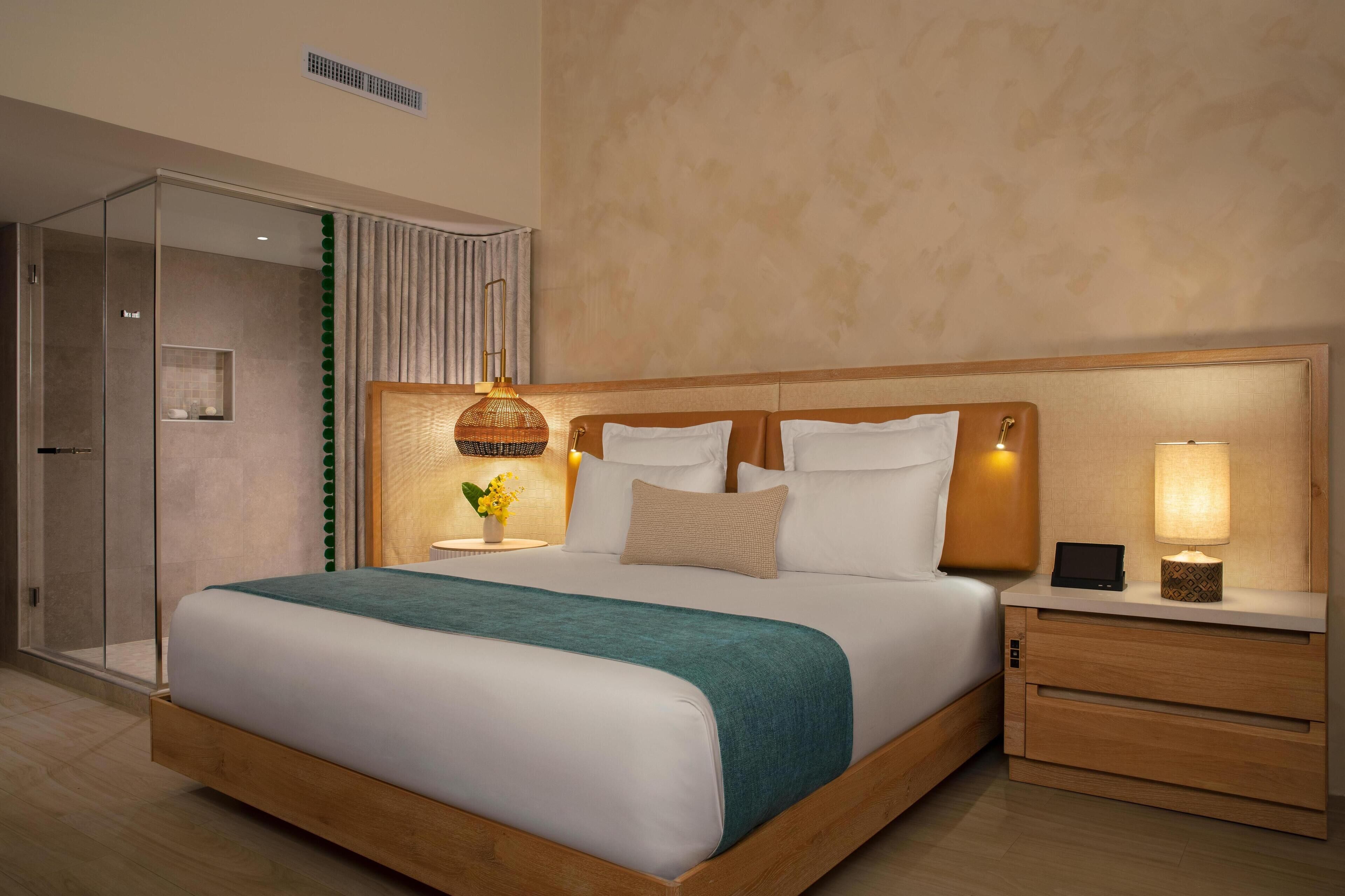 Una habitación de hotel moderna con una cama grande, mesitas de noche, una lámpara de noche y un box de ducha de vidrio.