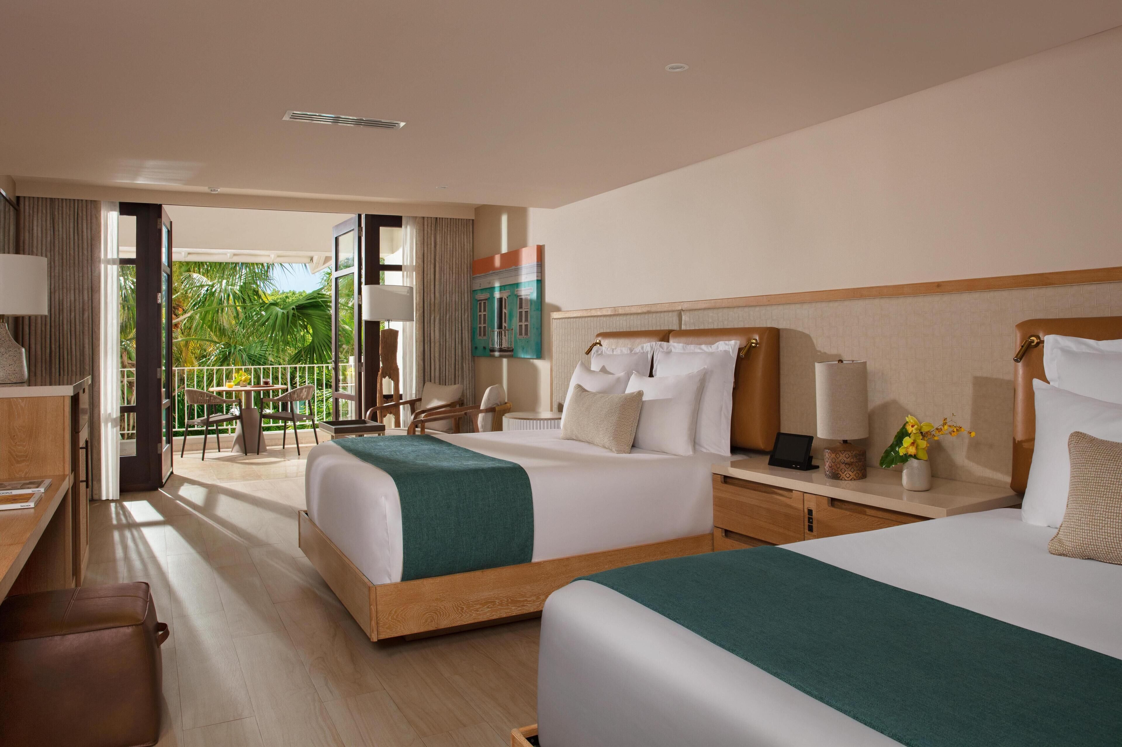 A hotel room with two beds, a desk, a chair, and a balcony with a view of palm trees.