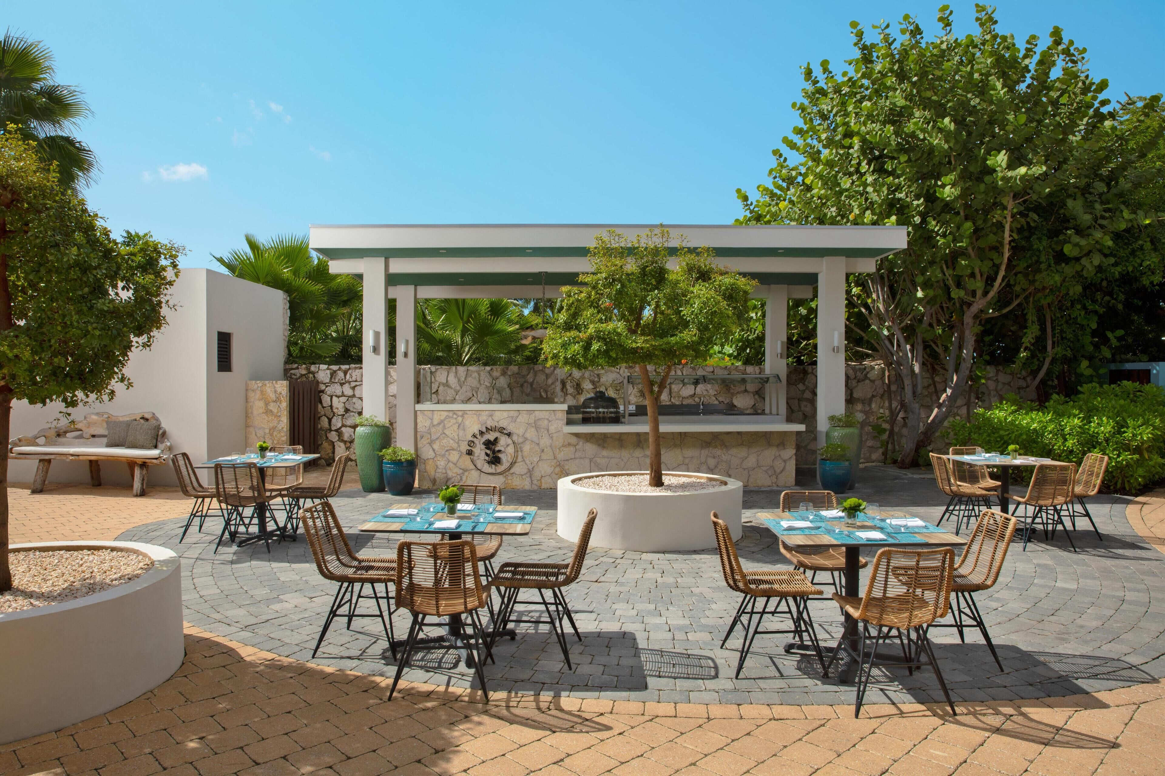 Un patio al aire libre con mesas y sillas, un muro de piedra y una pérgola con un árbol.