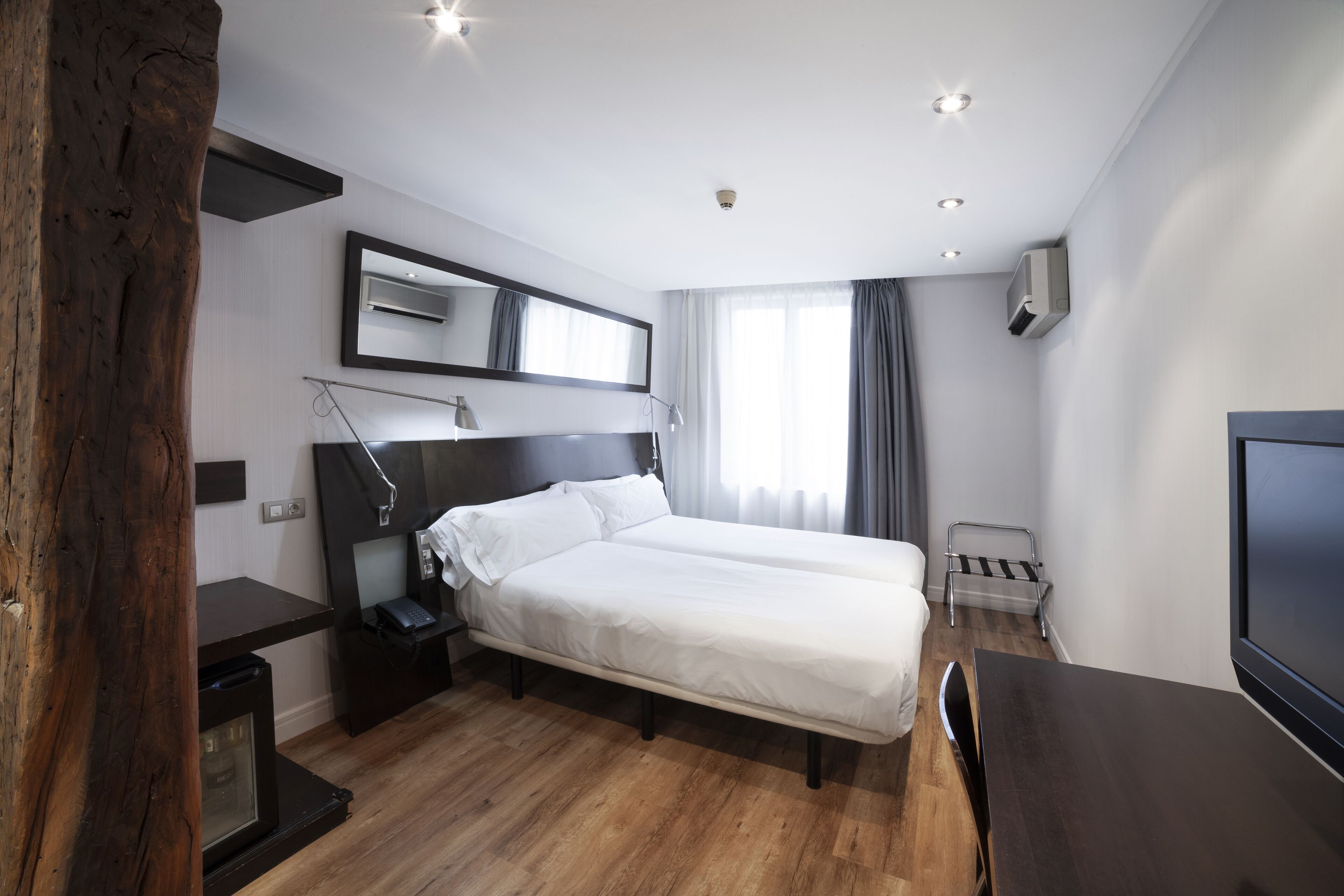 Una habitación de hotel moderna con piso de madera, una cama grande, un escritorio, una silla, un televisor y un espejo.
