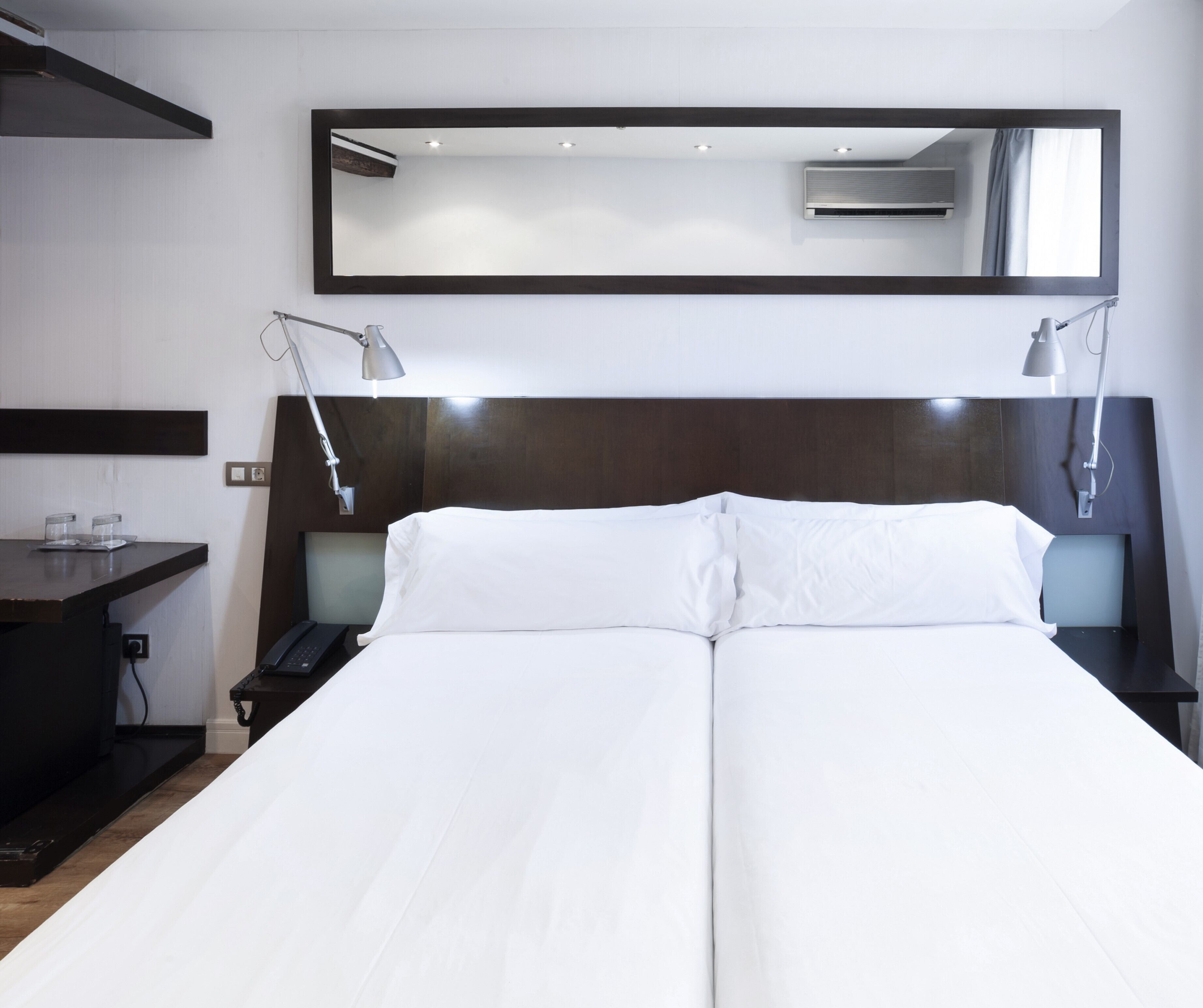 Un lit double avec du linge de lit blanc, une tête de lit sombre, deux tables de chevet avec des lampes, un bureau avec un téléphone et un grand miroir au-dessus du lit.