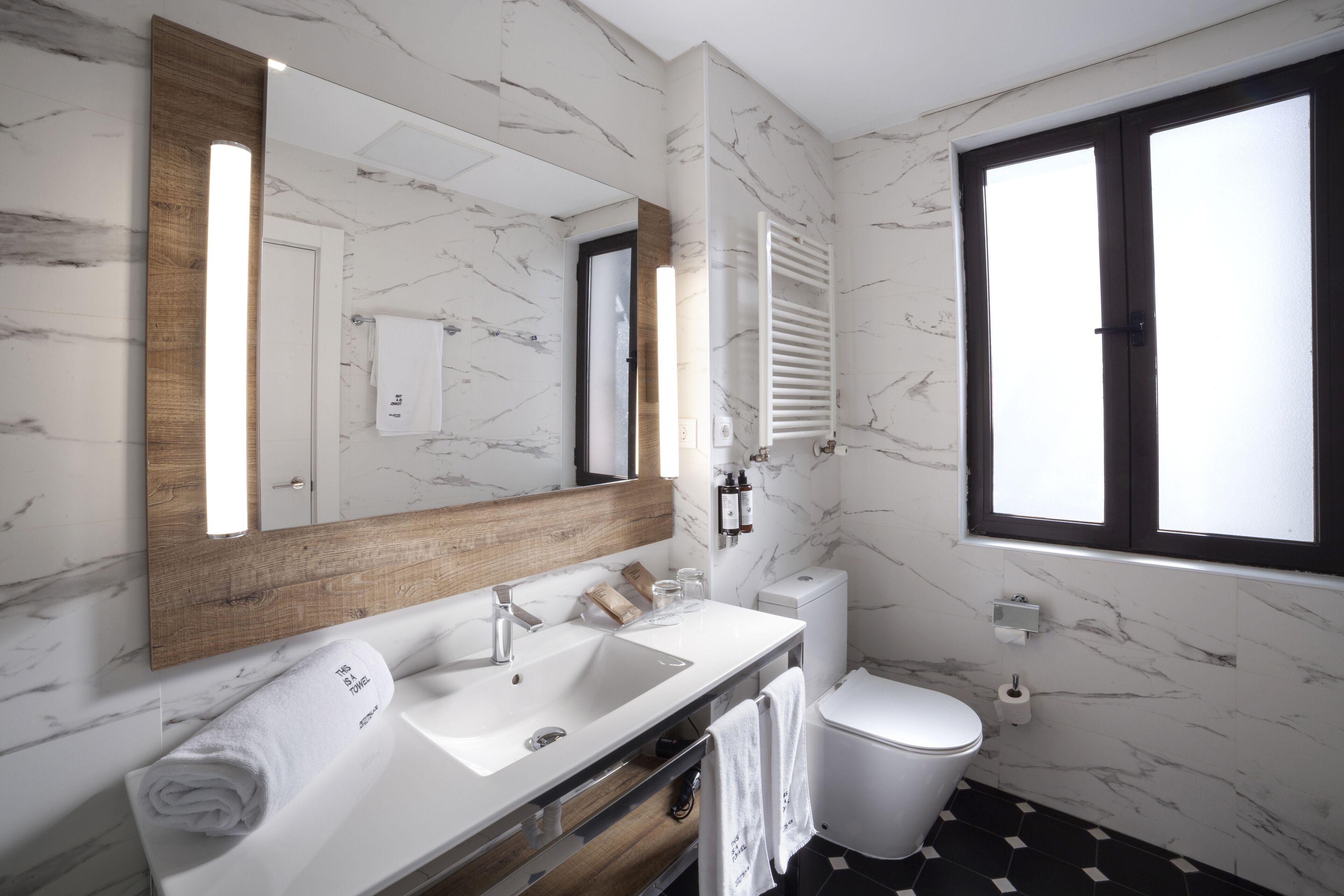 A modern bathroom with a marble backsplash, a wooden vanity, a large mirror, a toilet, and a window providing natural light.