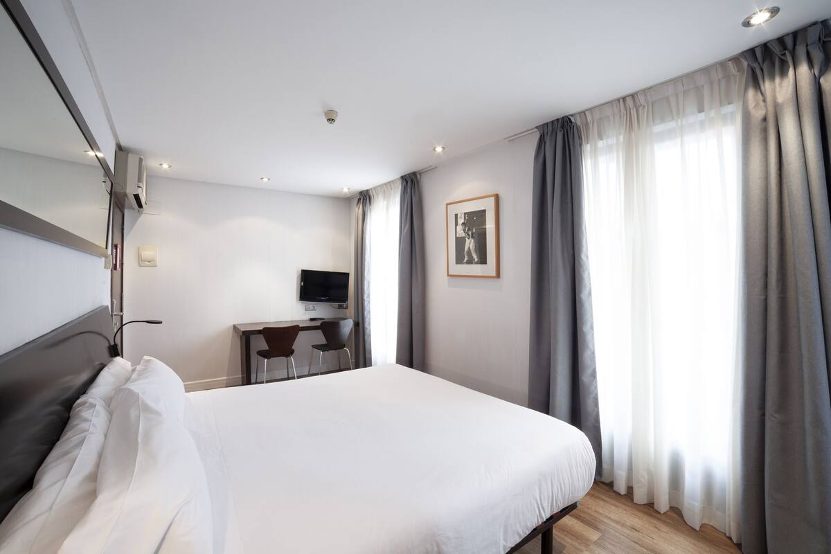 A modern hotel room with a large bed, a desk with a chair, a flat-screen TV, and a large window with curtains.