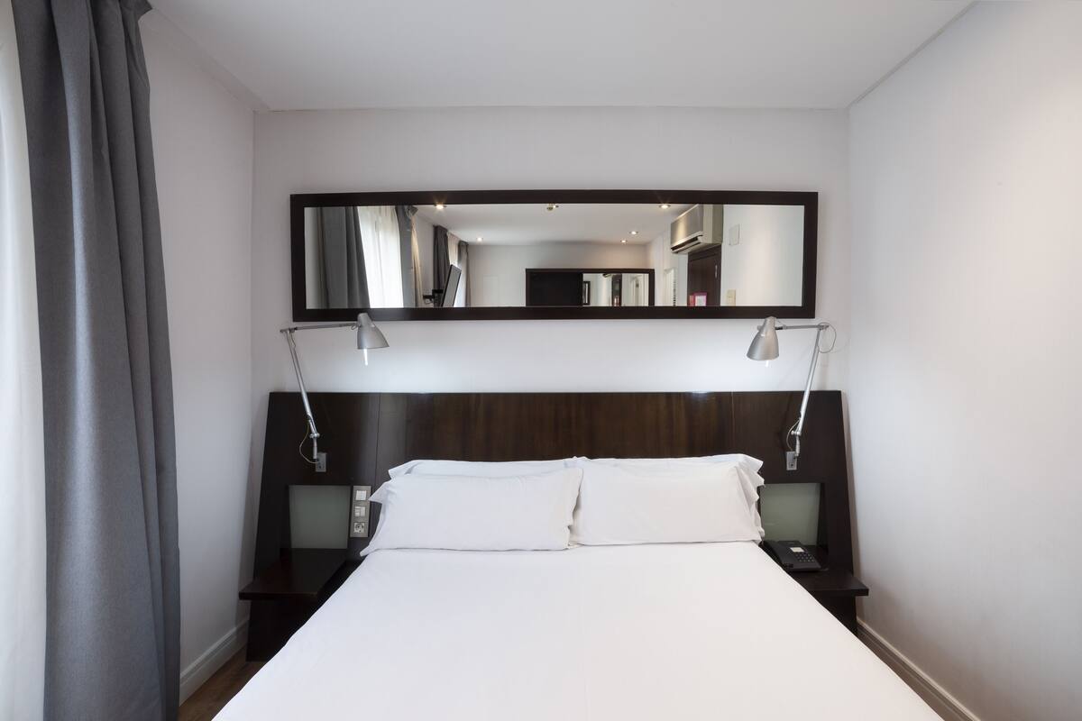 A hotel room with a large bed, a dark headboard, two bedside tables with lamps, and a large mirror above the bed.