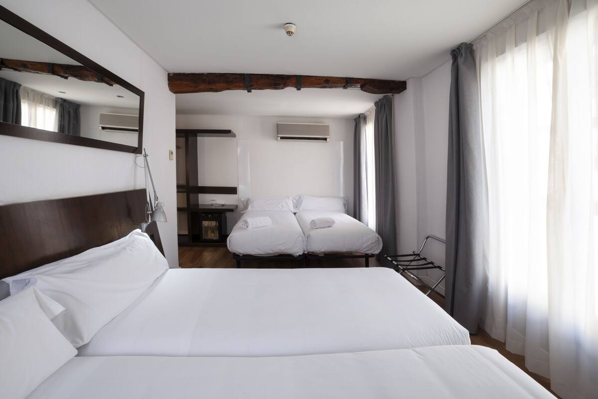 A hotel room with a large bed, a smaller bed, a mirror, an air conditioning unit, and a window with curtains.