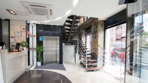 A modern hotel lobby with a reception desk, a staircase, and a view of the street outside. - Petit Palace Arana (Bilbao)