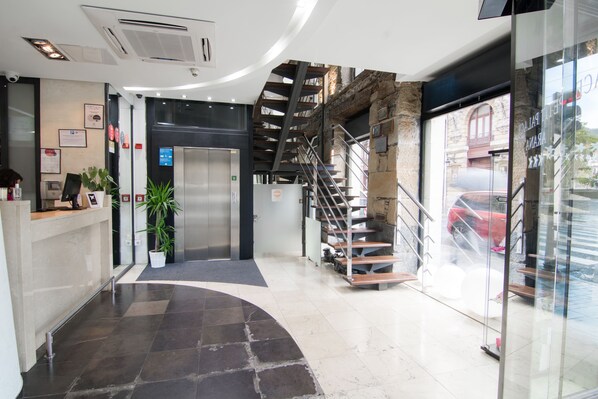A modern hotel lobby with a reception desk, a staircase, and a view of the street outside. - Petit Palace Arana (Bilbao)