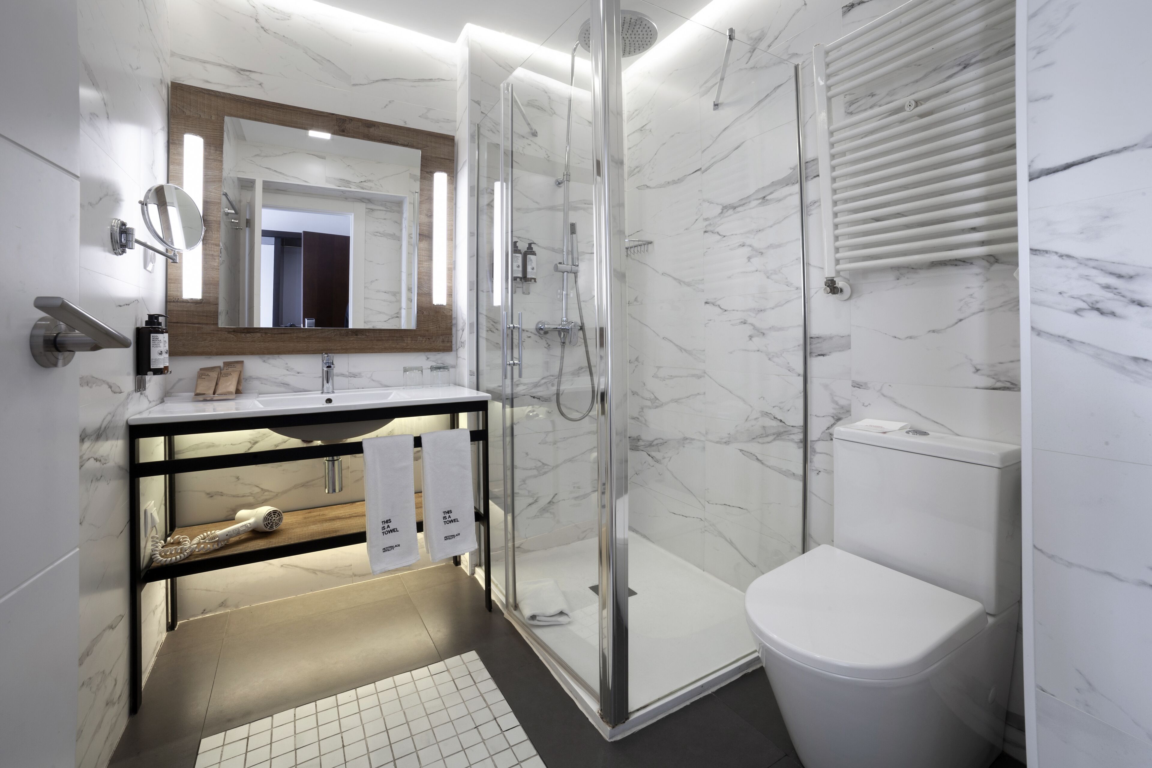 Un baño moderno con pared de mármol, cabina de ducha de vidrio, inodoro blanco y tocador con espejo y toallero.