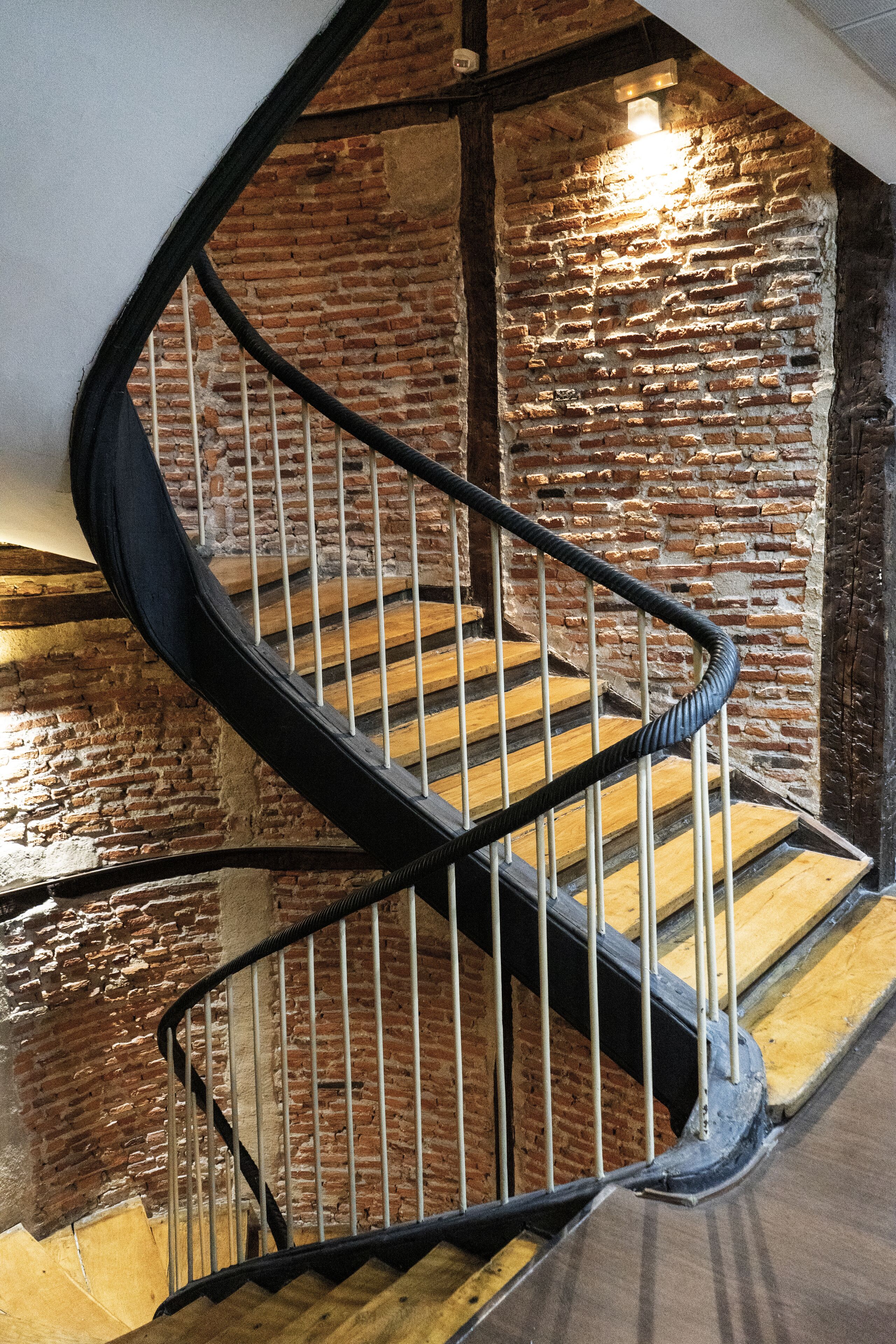 A spiral staircase with wooden steps and metal railings, leading to a brick wall with a light fixture.