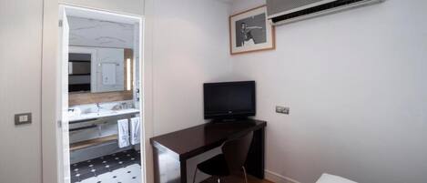A small, modern room with a wooden desk, a black chair, a flat-screen TV, and an air conditioning unit mounted on the wall.