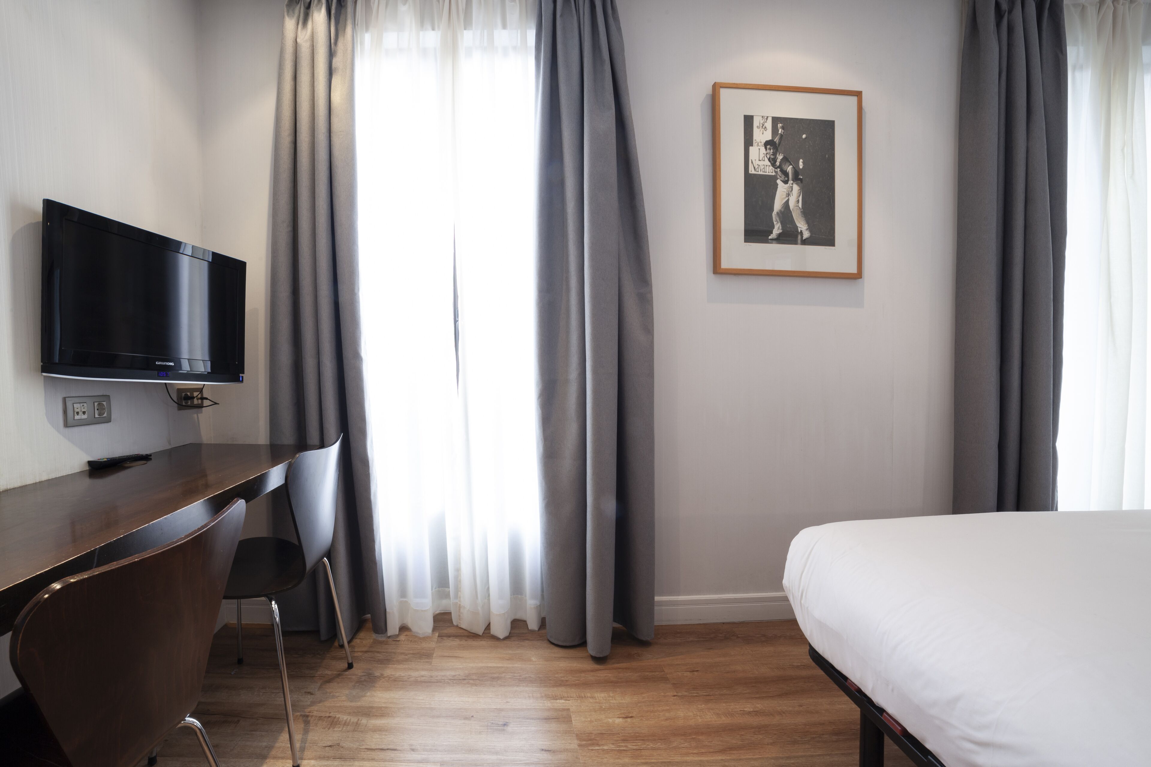 Hotellihuone, jossa on puinen työpöytä, litteänäyttöinen televisio, sänky ja seinällä oleva kehystetty kuva.