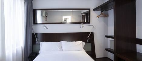 A hotel room with a bed, bedside lamps, a large mirror, a wooden wardrobe, and a hanging coat rack.
