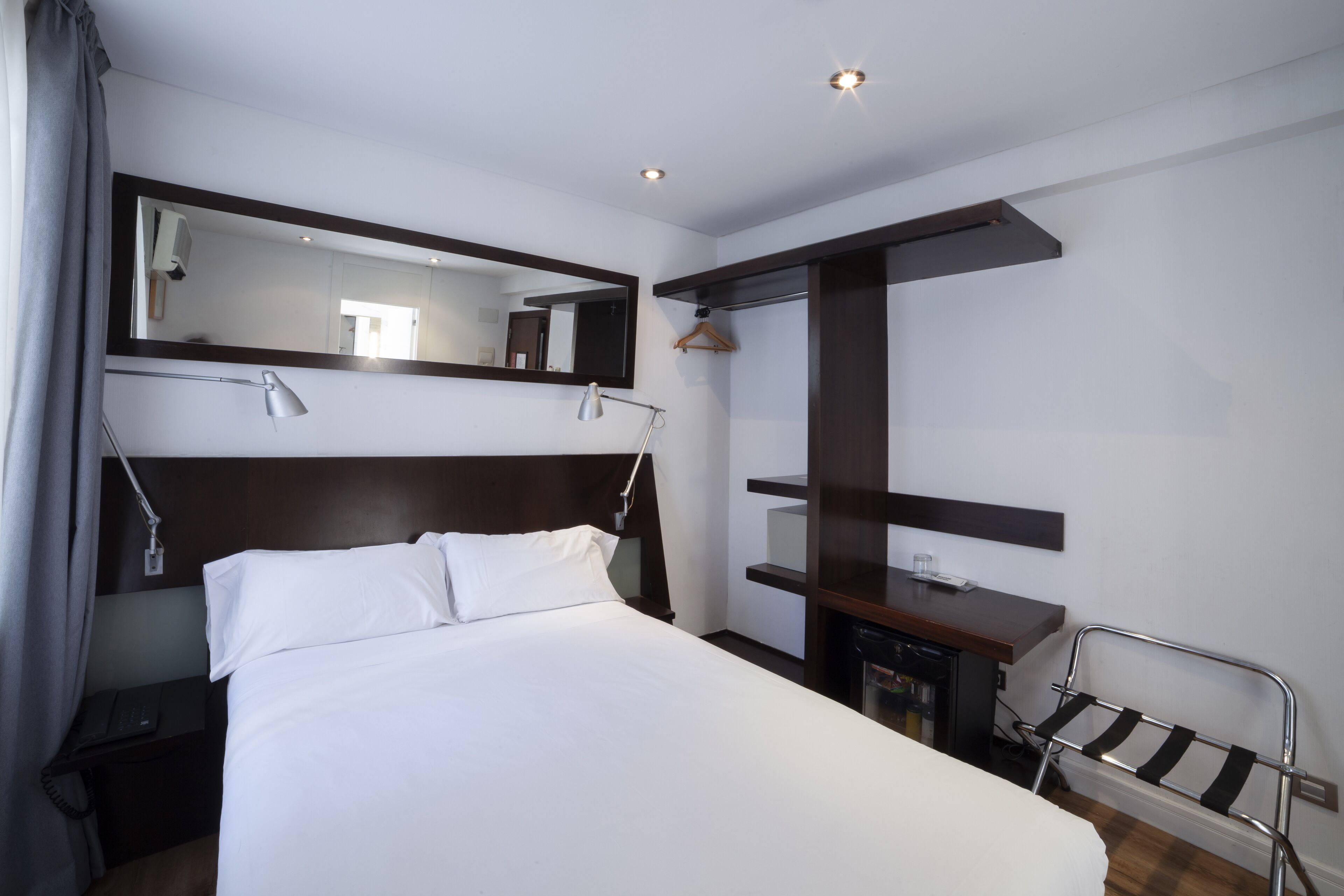 A hotel room with a bed, bedside table, lamp, mirror, and a small desk with a chair.