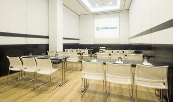 Ein Konferenzraum mit einer großen Leinwand, auf der das LUNION Hotels Logo zu sehen ist, weißen Stühlen und Tischen, die für Besprechungen bereitstehen.