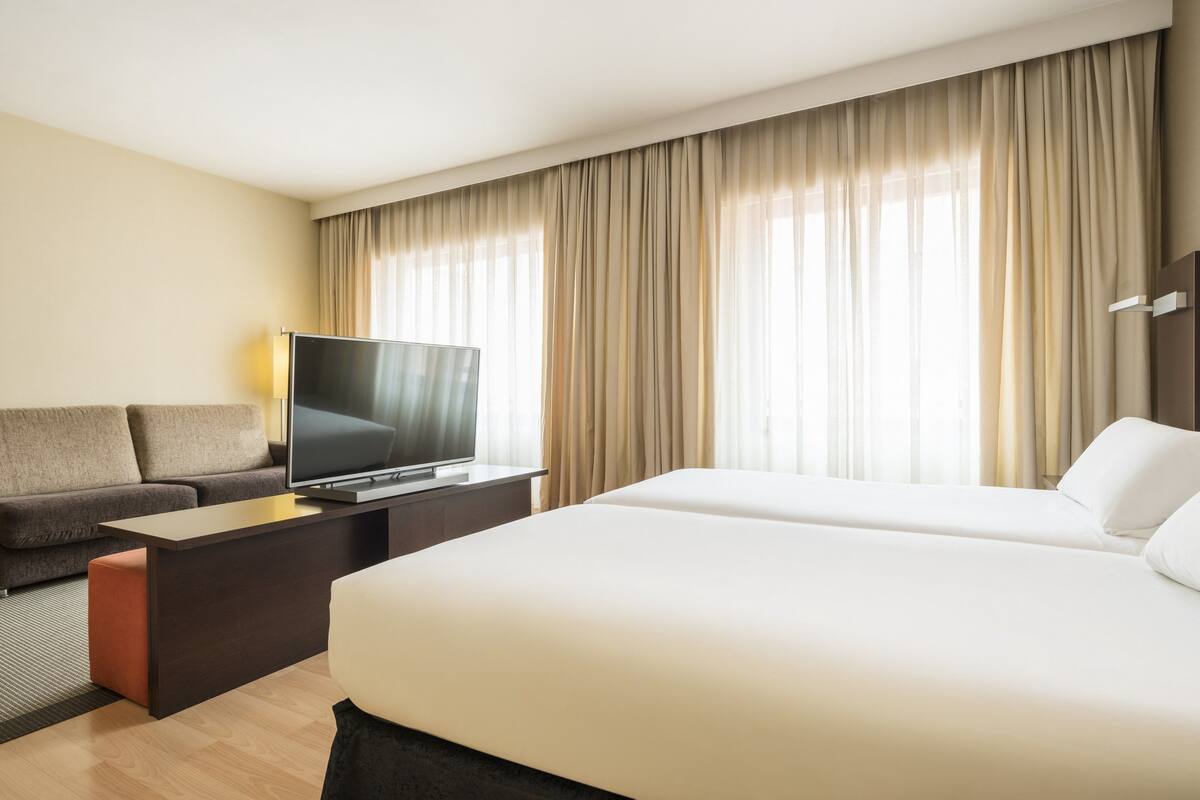A hotel room with a large bed, a sofa, a TV on a stand, and a window with curtains.