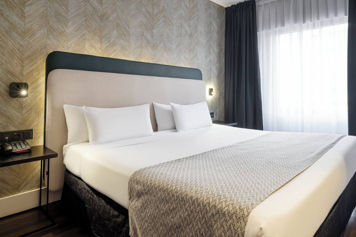 A hotel room with a large bed, a bedside table with a phone, and a wall-mounted light.