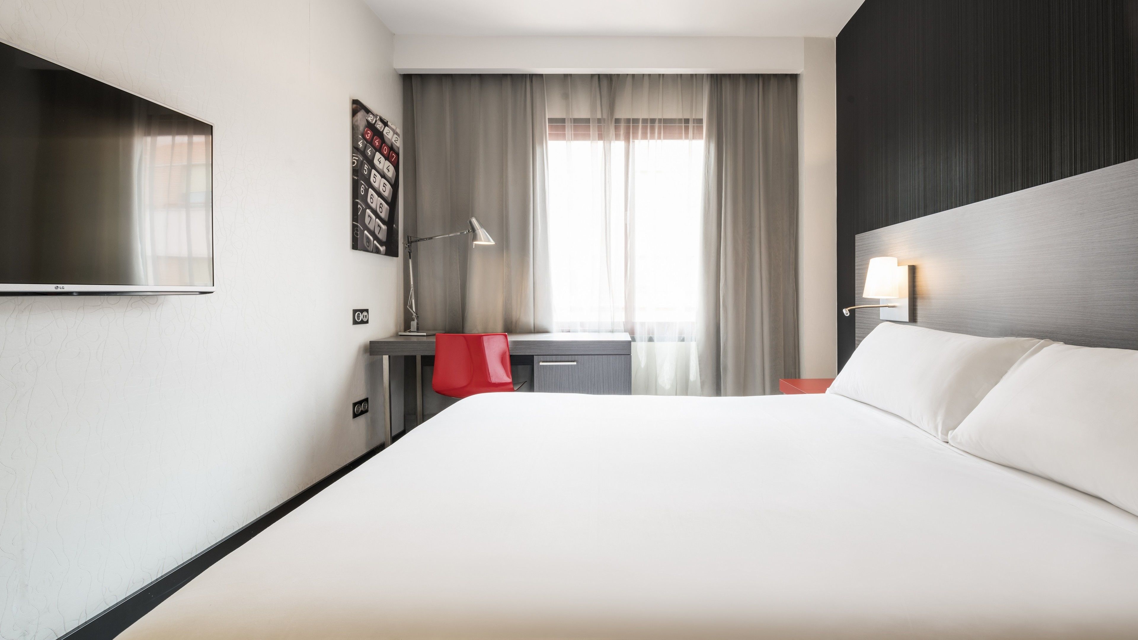Een hotelkamer met een groot bed, een bureau met stoel, een aan de muur bevestigde televisie en een raam met gordijnen.