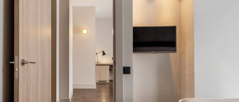 A modern hotel room with a wooden door, a bed with a patterned bedspread, a mounted flat-screen TV, and a hallway leading to a bathroom.