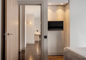Ein modernes Hotelzimmer mit Holztür, einem Bett mit gemustertem Überwurf, einem wandmontierten Flachbildfernseher und einem zur Toilette führenden Flur.