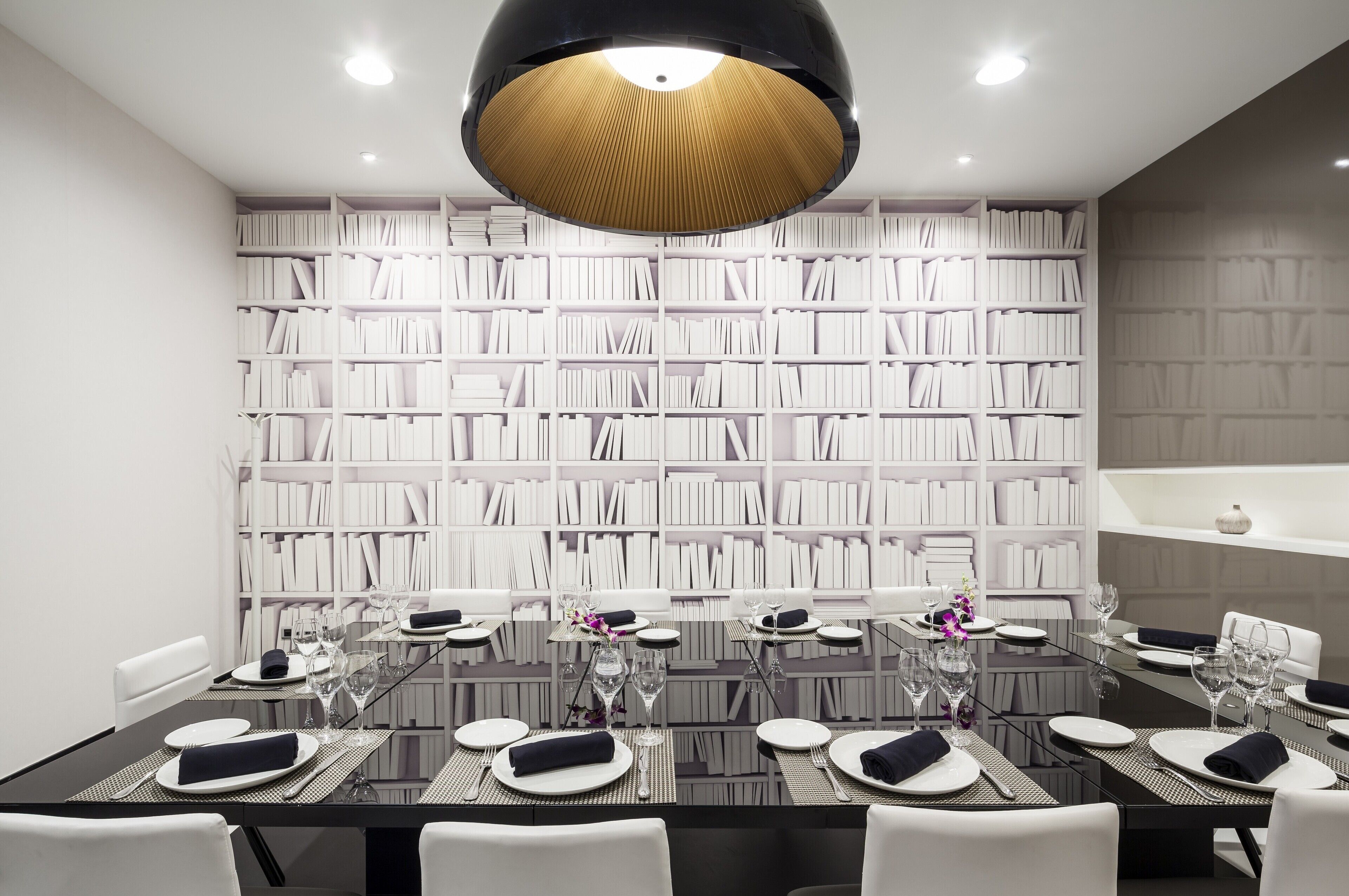 Comedor con una mesa grande dispuesta para seis personas, rodeada de sillas blancas, y una estantería al fondo.