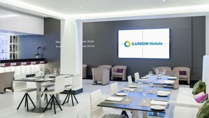 A modern hotel lobby with a large screen displaying the hotel name, multiple dining tables set for guests, and comfortable seating areas. - Hotel ILUNION Suites Madrid (Madrid)